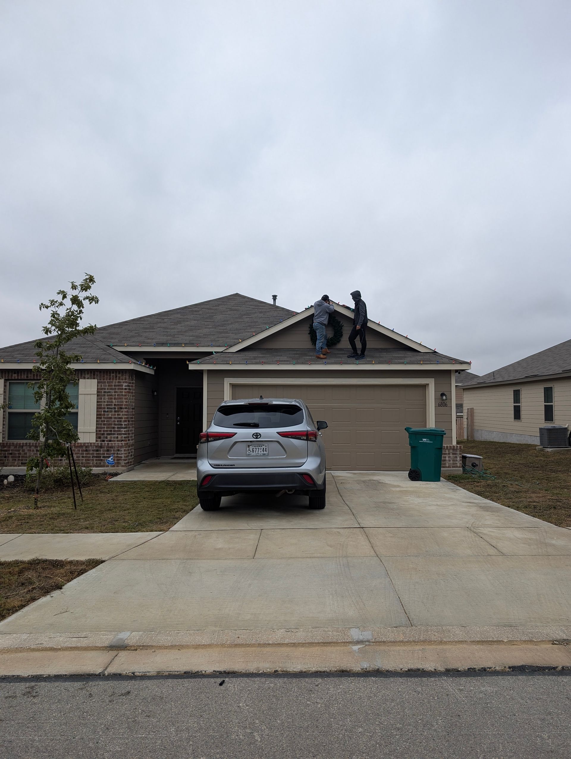 Two workers on a house roof, gray car in driveway, overcast sky.
