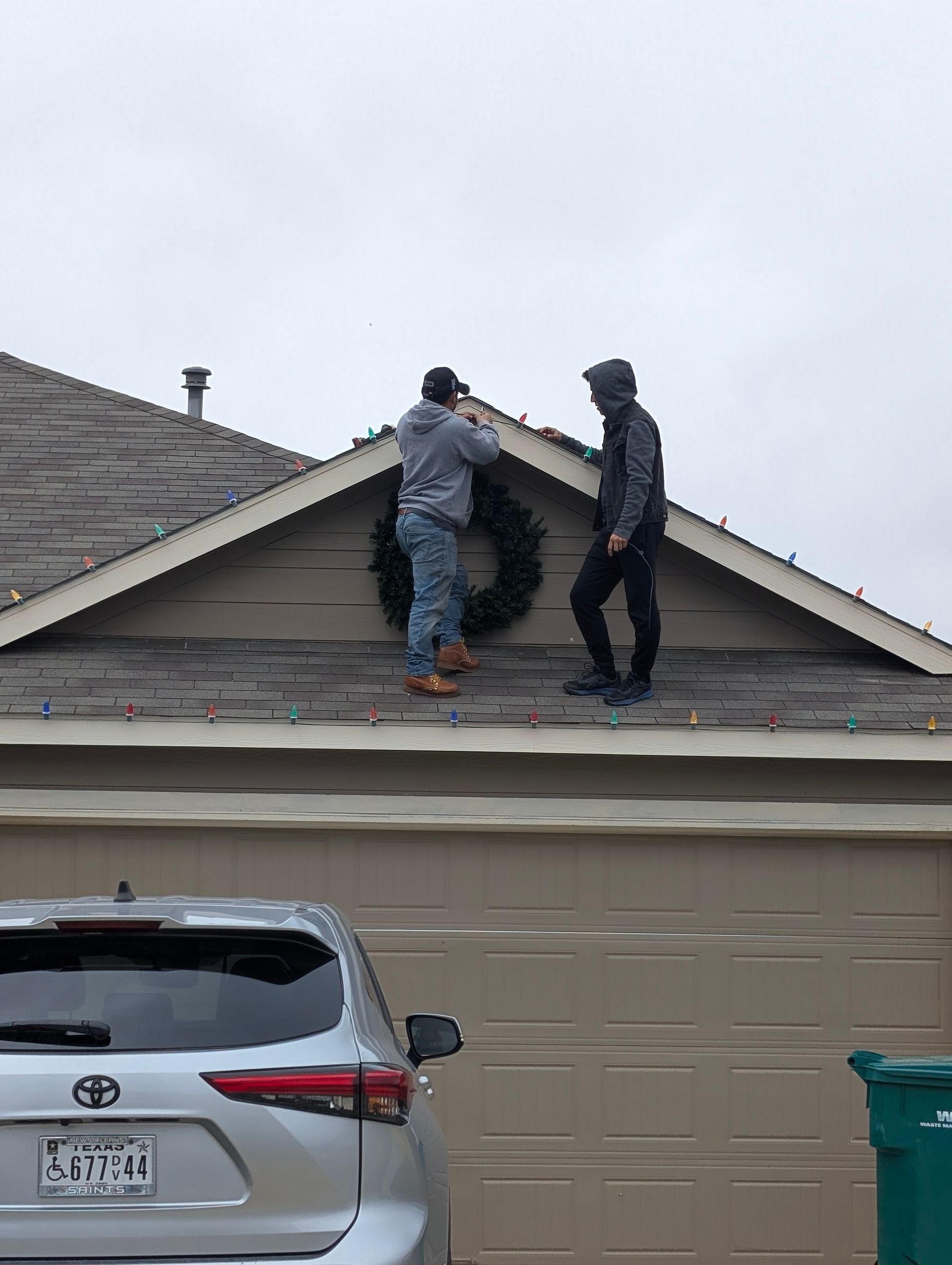 Two people hanging a wreath on a house roof. Overcast sky, beige siding, a car parked below.
