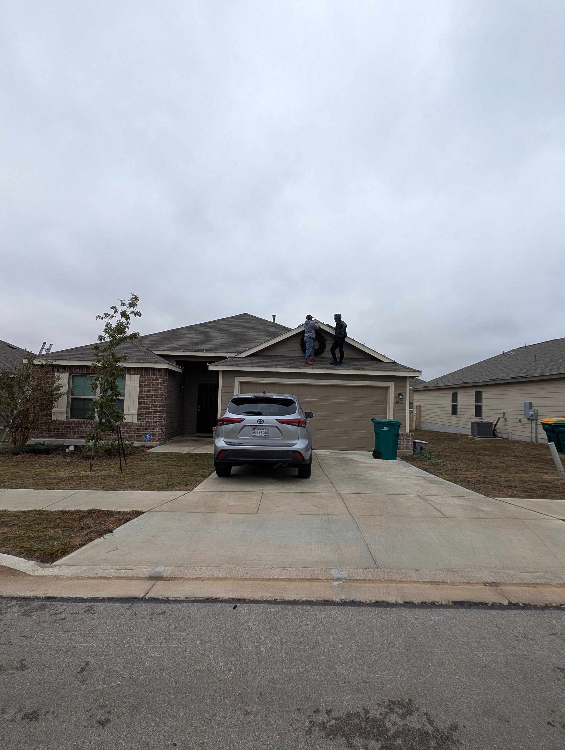 House with two people on the roof; cars in the driveway. Overcast day in a suburban setting.
