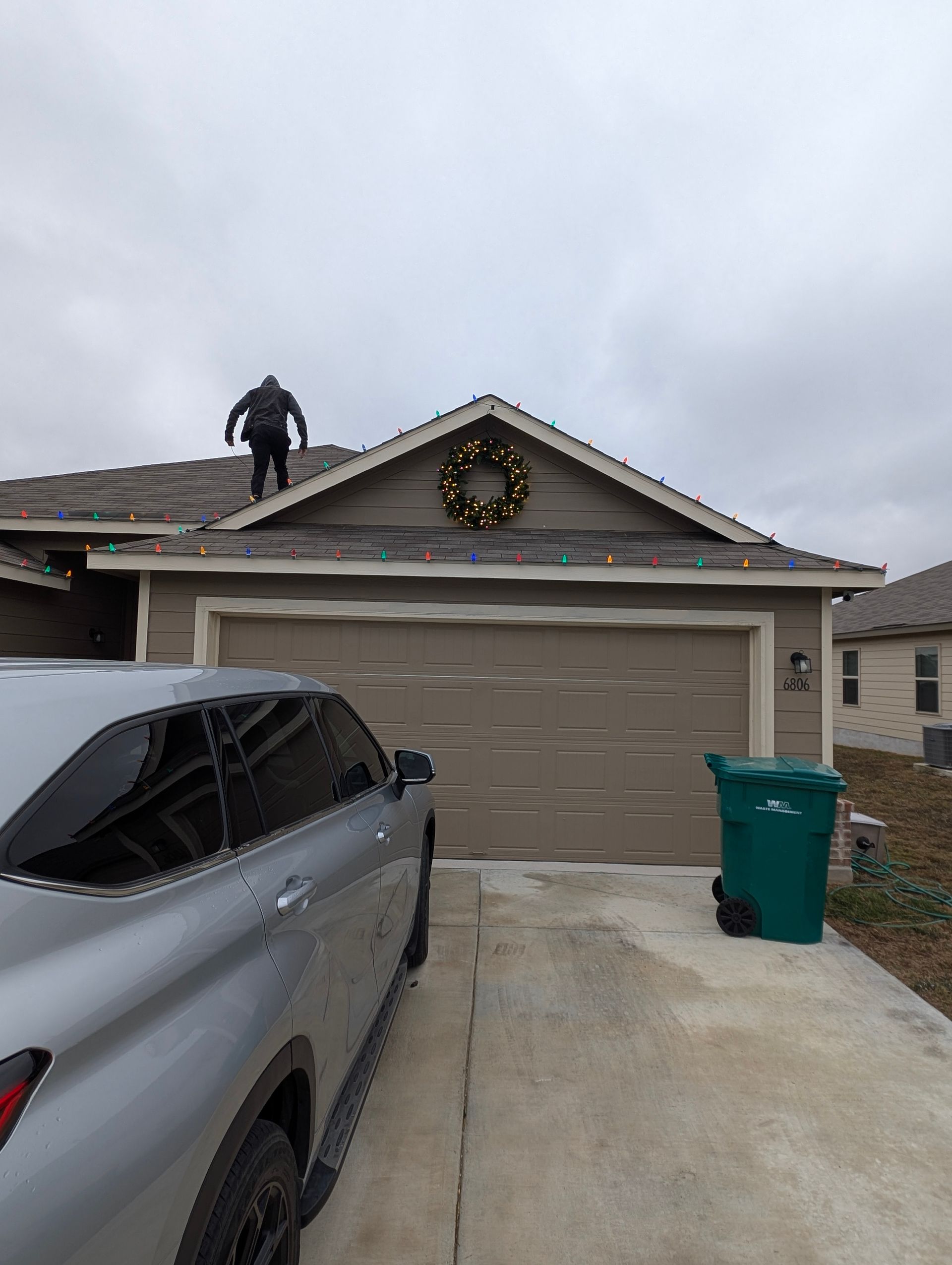 Man on roof, hanging wreath. Garage below, car parked in front. Cloudy day.