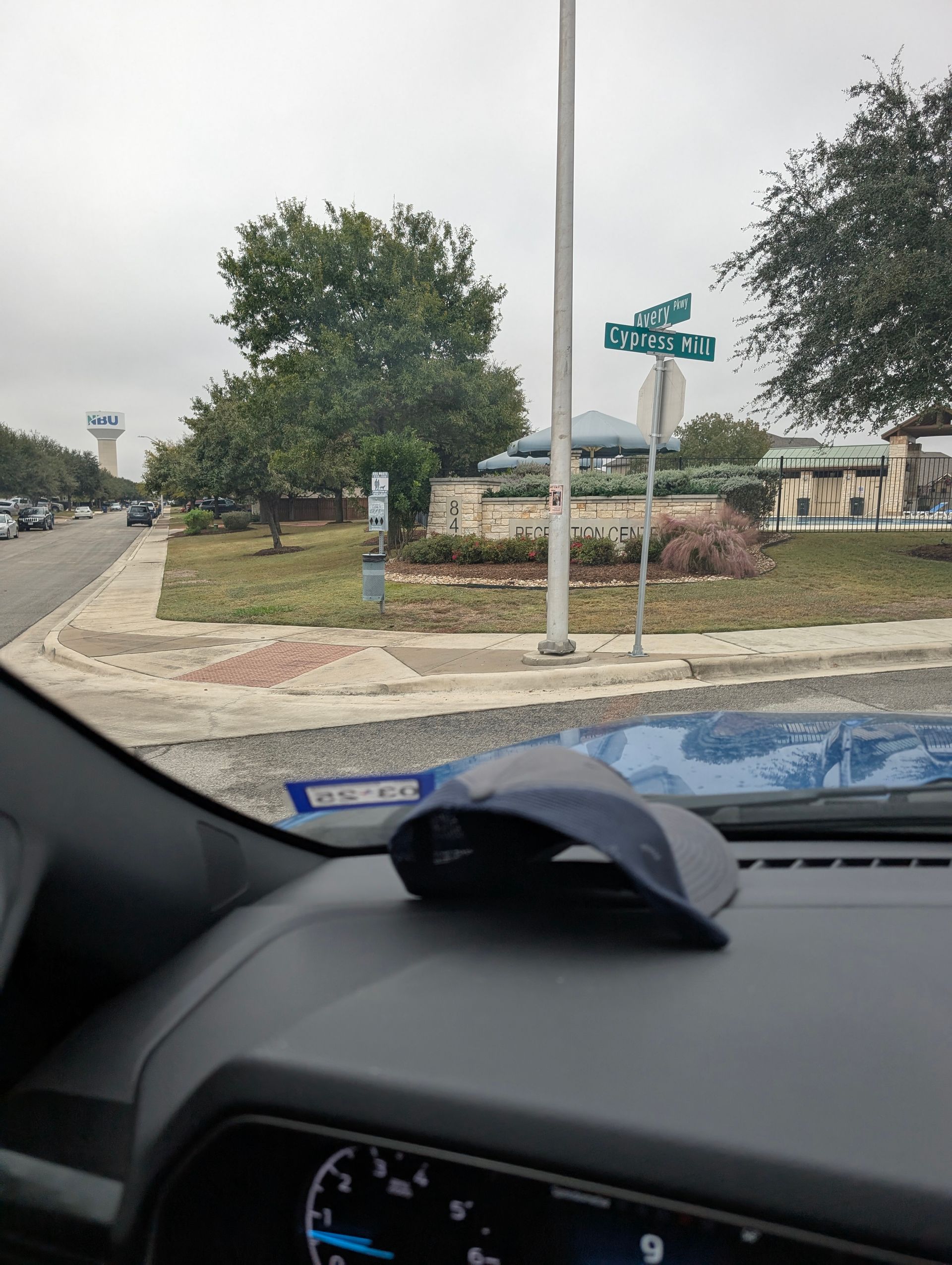 View from a car dashboard. Street corner with signs, a tree, and a cloudy sky. A blue hat rests on the dash.