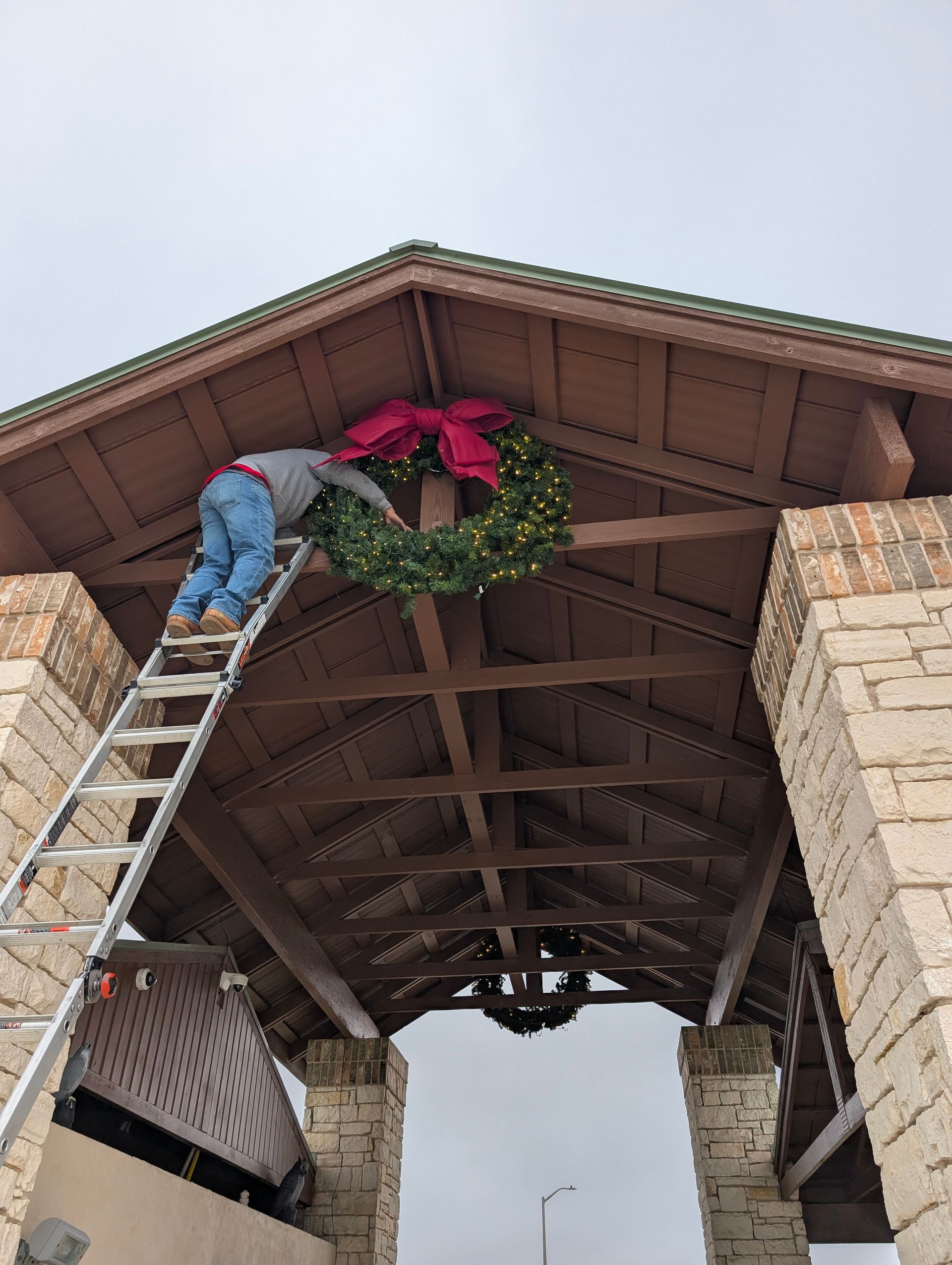 Person on a ladder hanging a Christmas wreath with a red bow on a wooden structure.