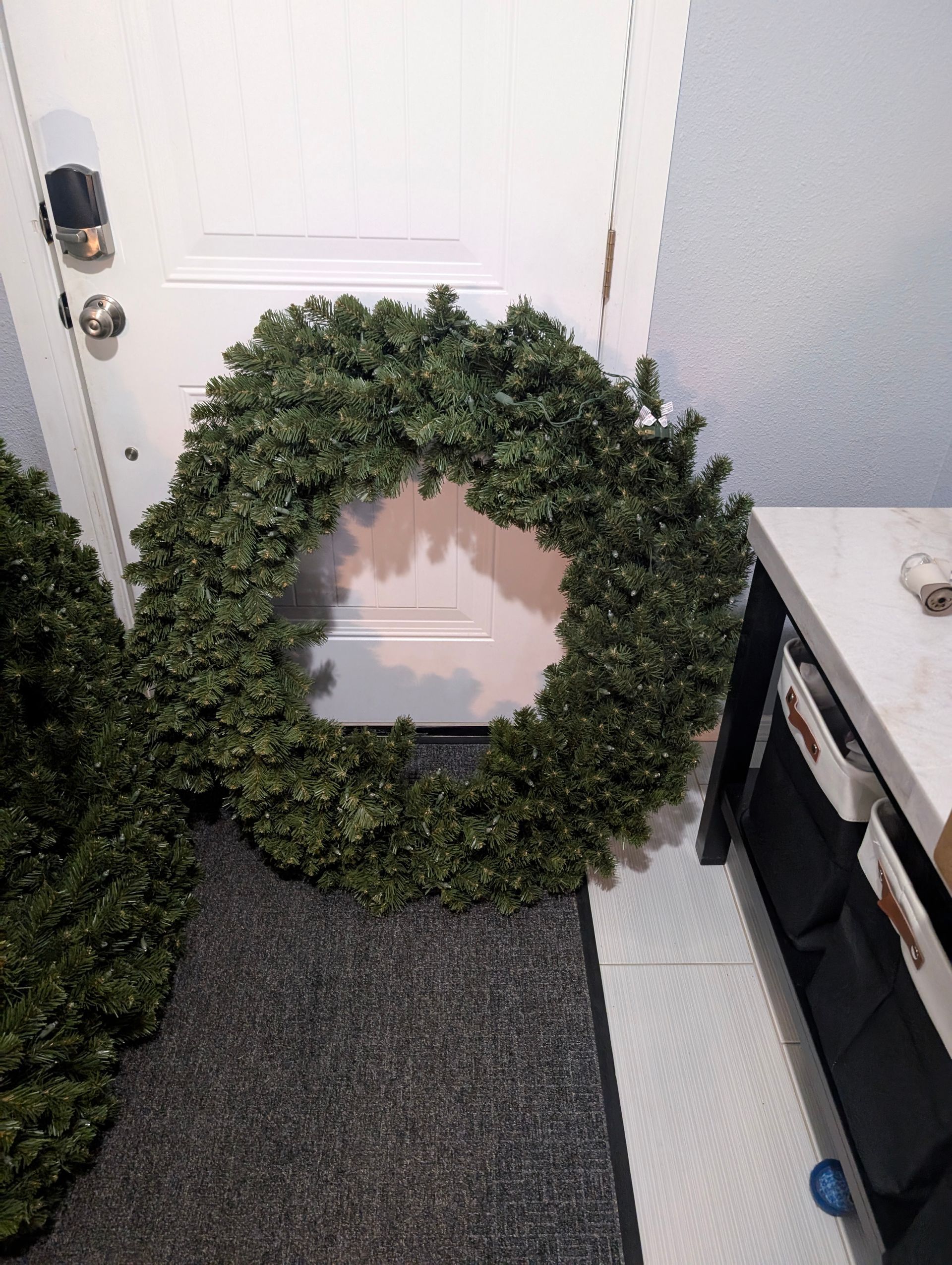 Green wreath resting against a white door, on a dark gray mat, inside a home.
