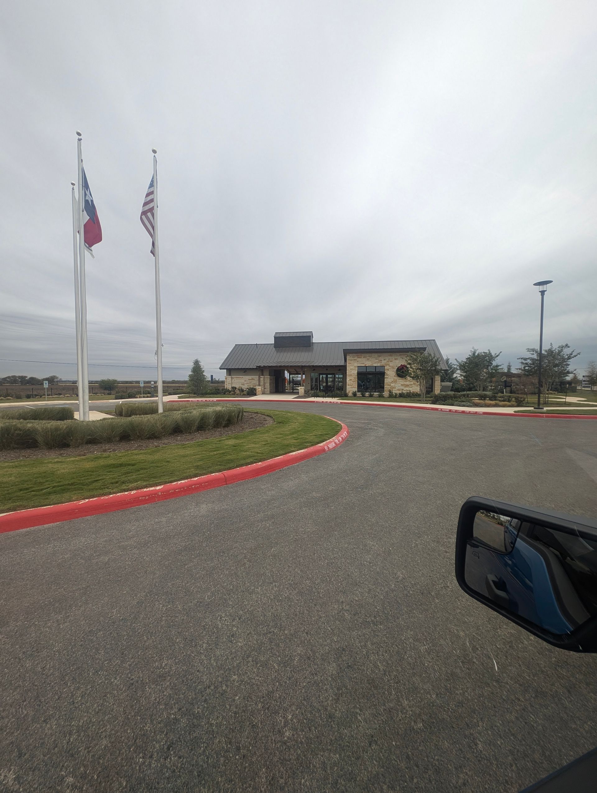 Building with flags, gray sky, and asphalt driveway, taken from inside a vehicle.