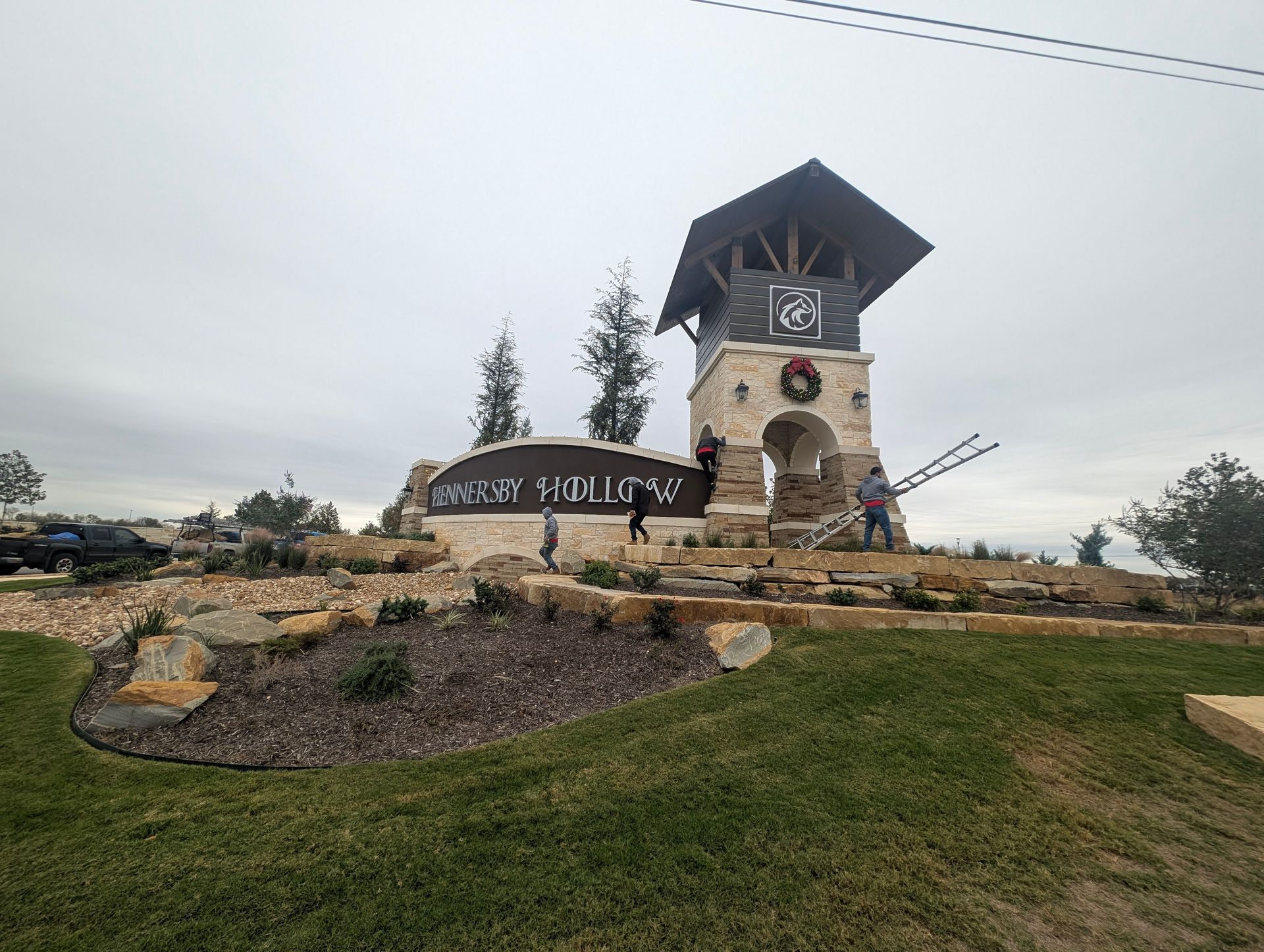 Entry sign for Sanctuary Point with clock tower, wreath, landscaping and people under overcast sky.