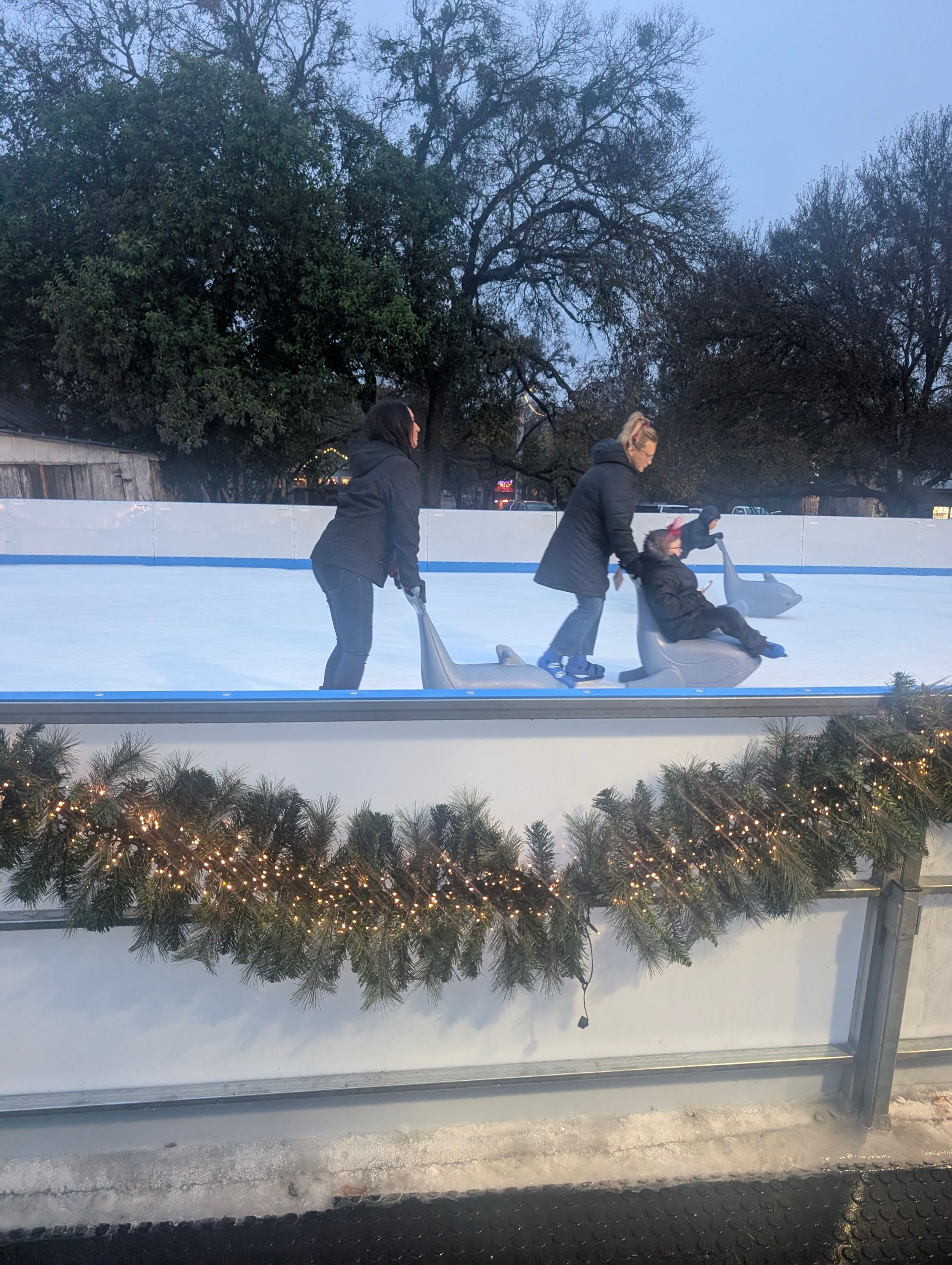 People ice skating on a rink, one person falls. Outdoors, green trees in the background.