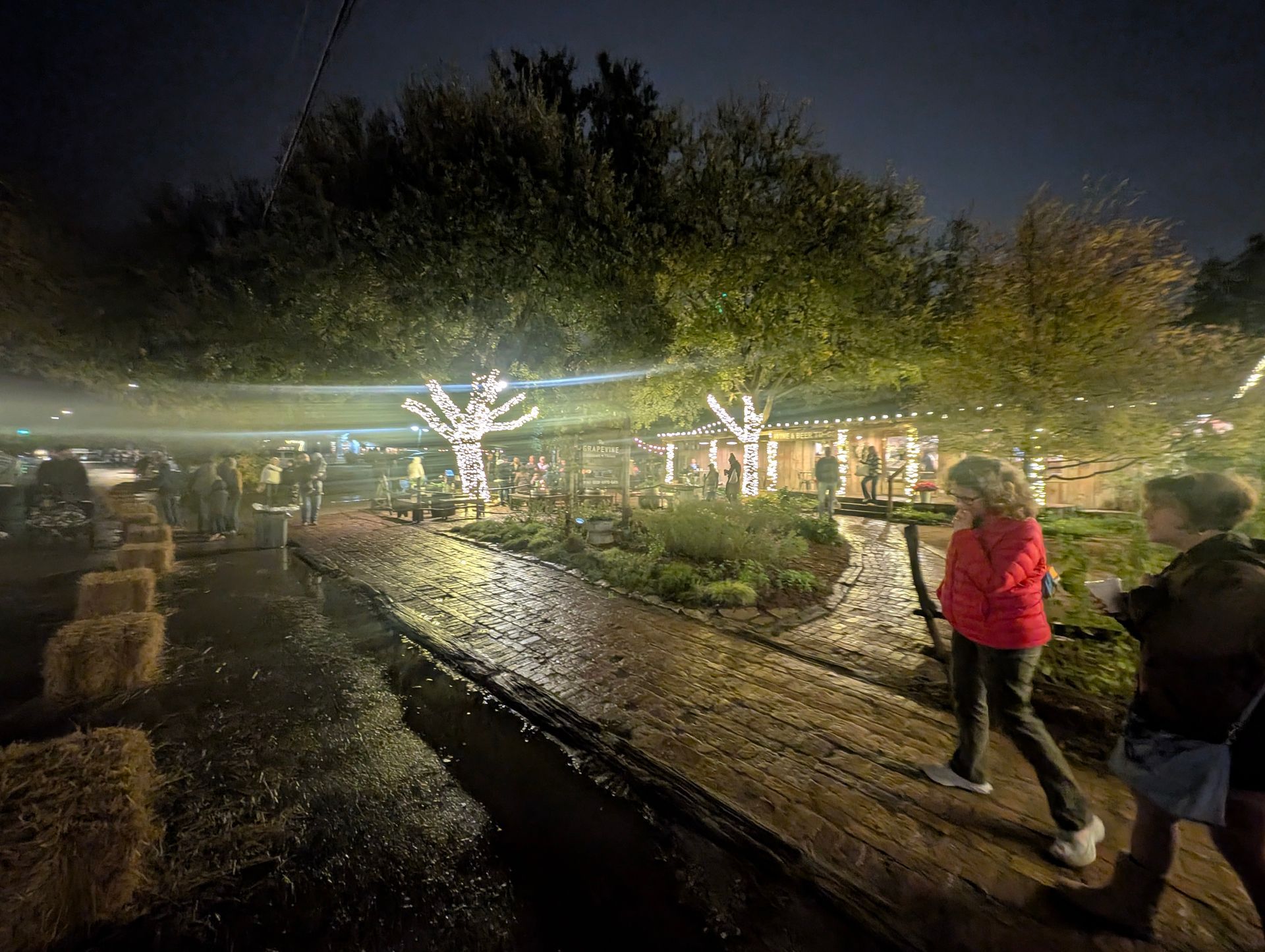 Nighttime scene: People walking on a wooden path, lit trees, and hay bales.