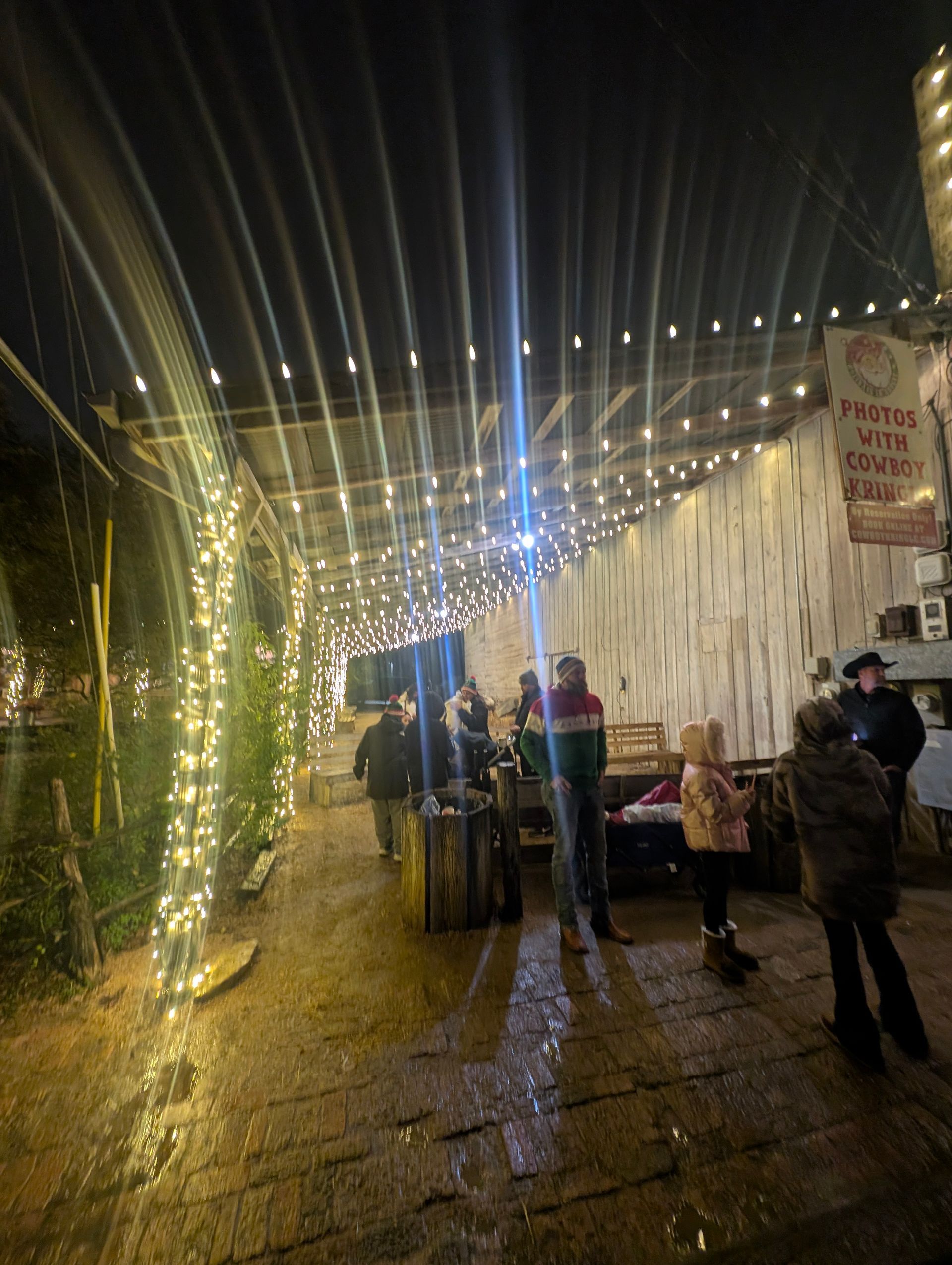 People gather under string lights at night. A festive market setting with a sign visible.