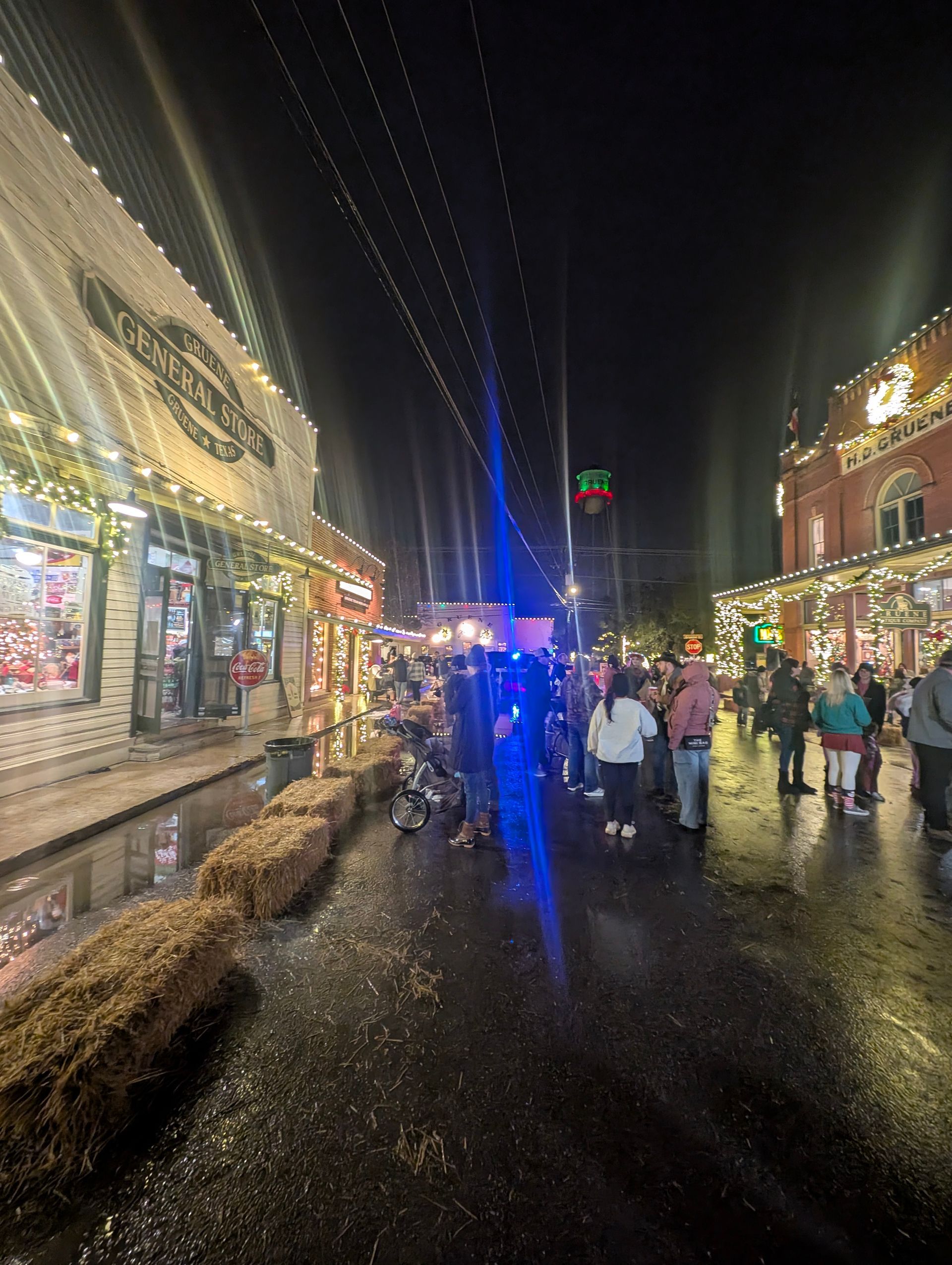 Nighttime street scene with storefronts and people, decorated with Christmas lights.