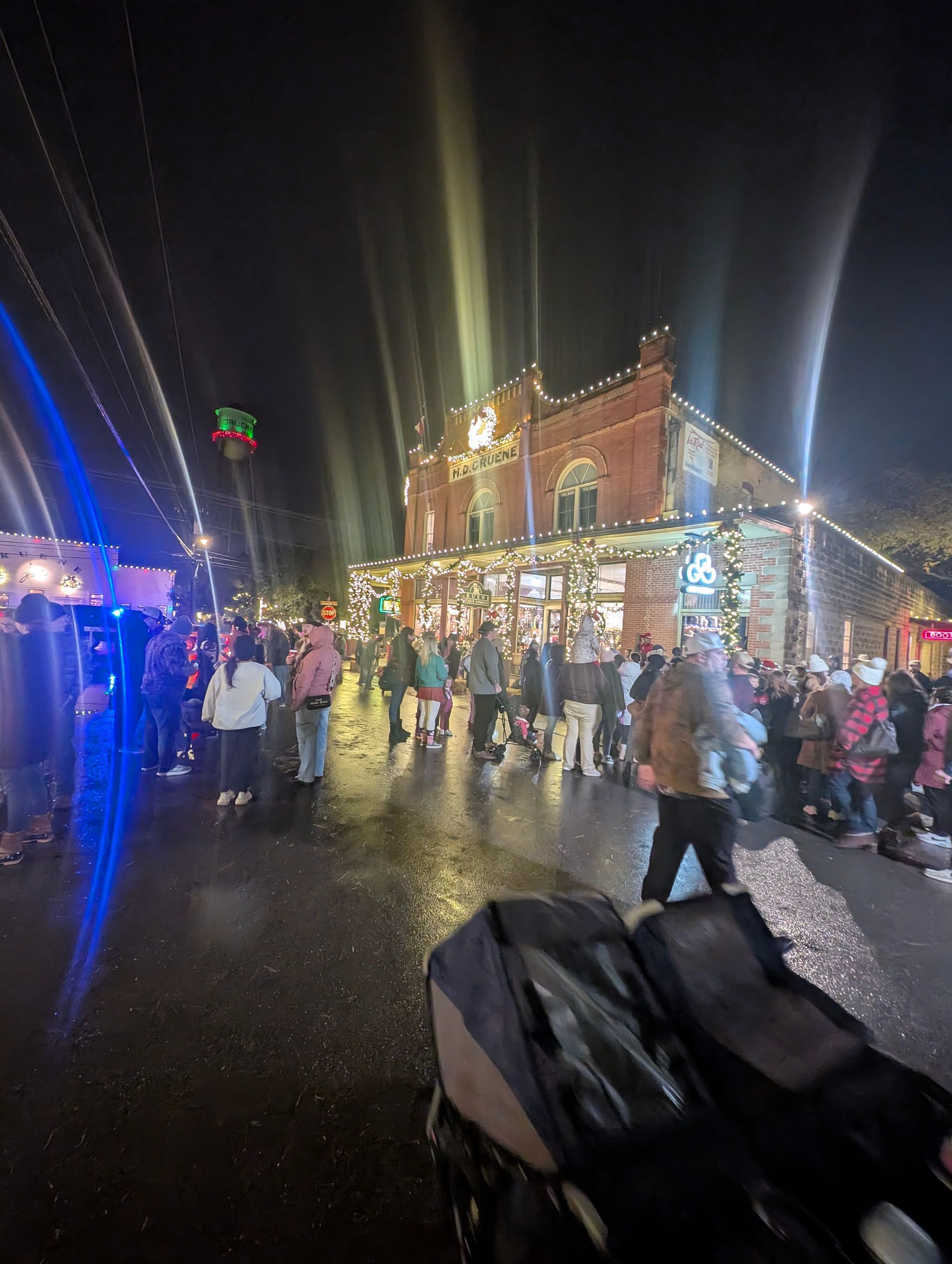 Crowd gathered on a street at night, lit by holiday lights, storefront, and people.