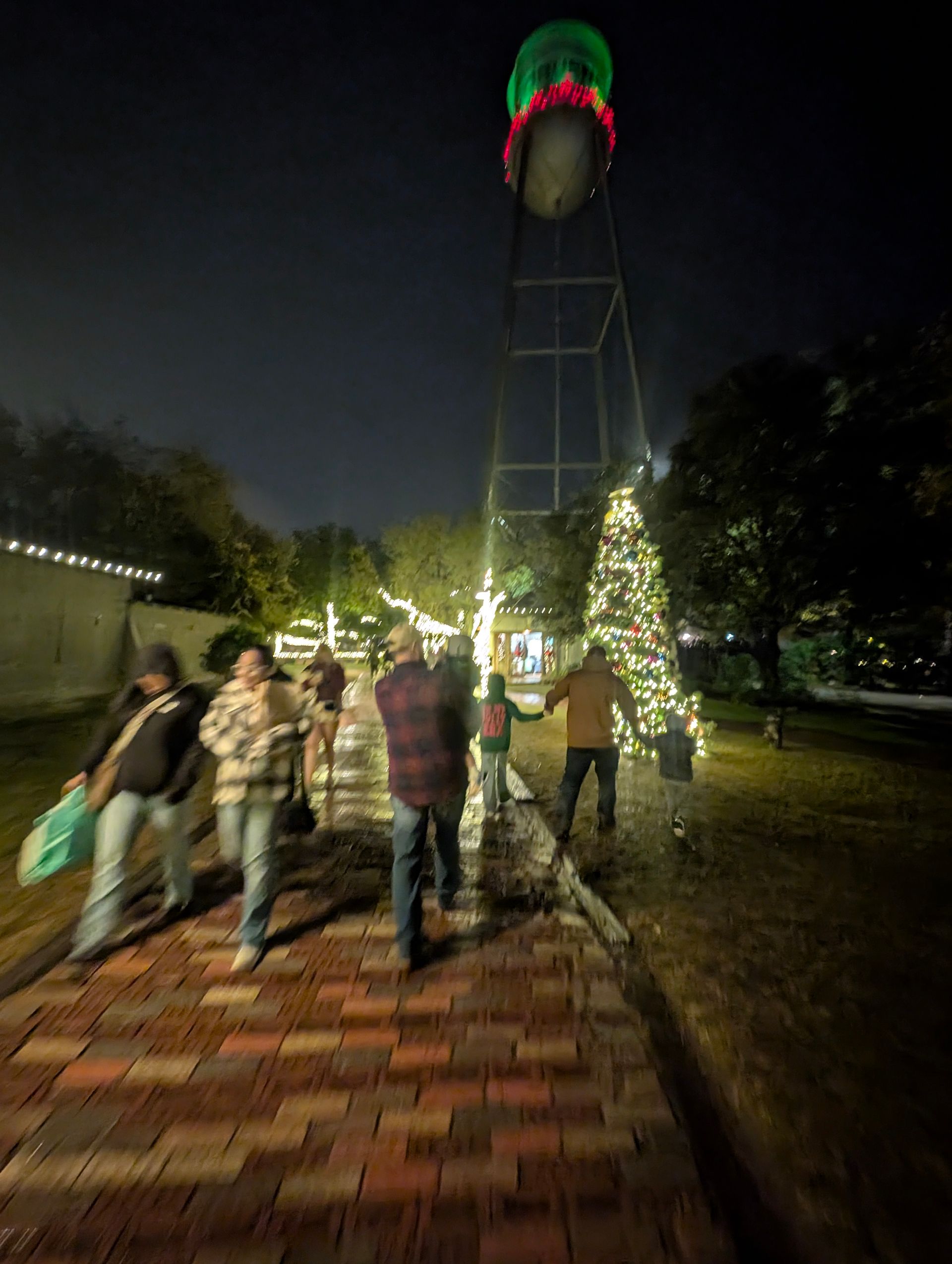People walking along a brick path at night with Christmas lights; a decorated water tower is visible in the background.