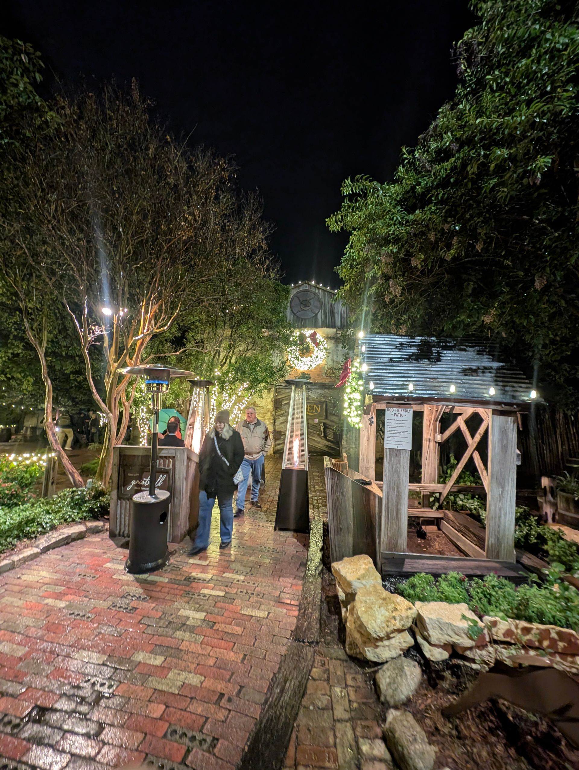 Night scene: People walking along a brick path lined with trees and lighted decor. Wooden structures and heaters are present.
