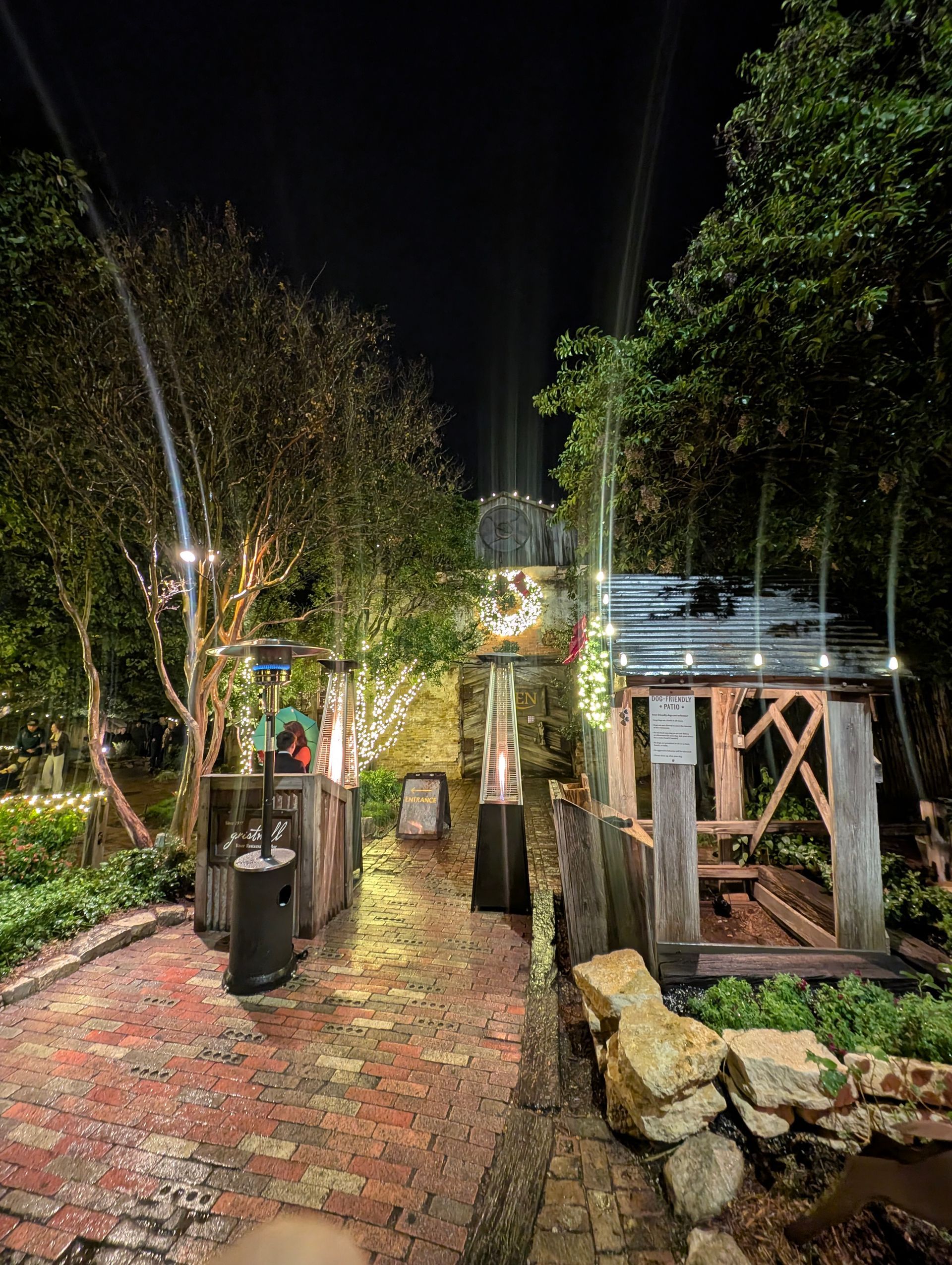 Night scene: brick path through a garden lit by string lights, outdoor heaters, and a wooden structure on the right.