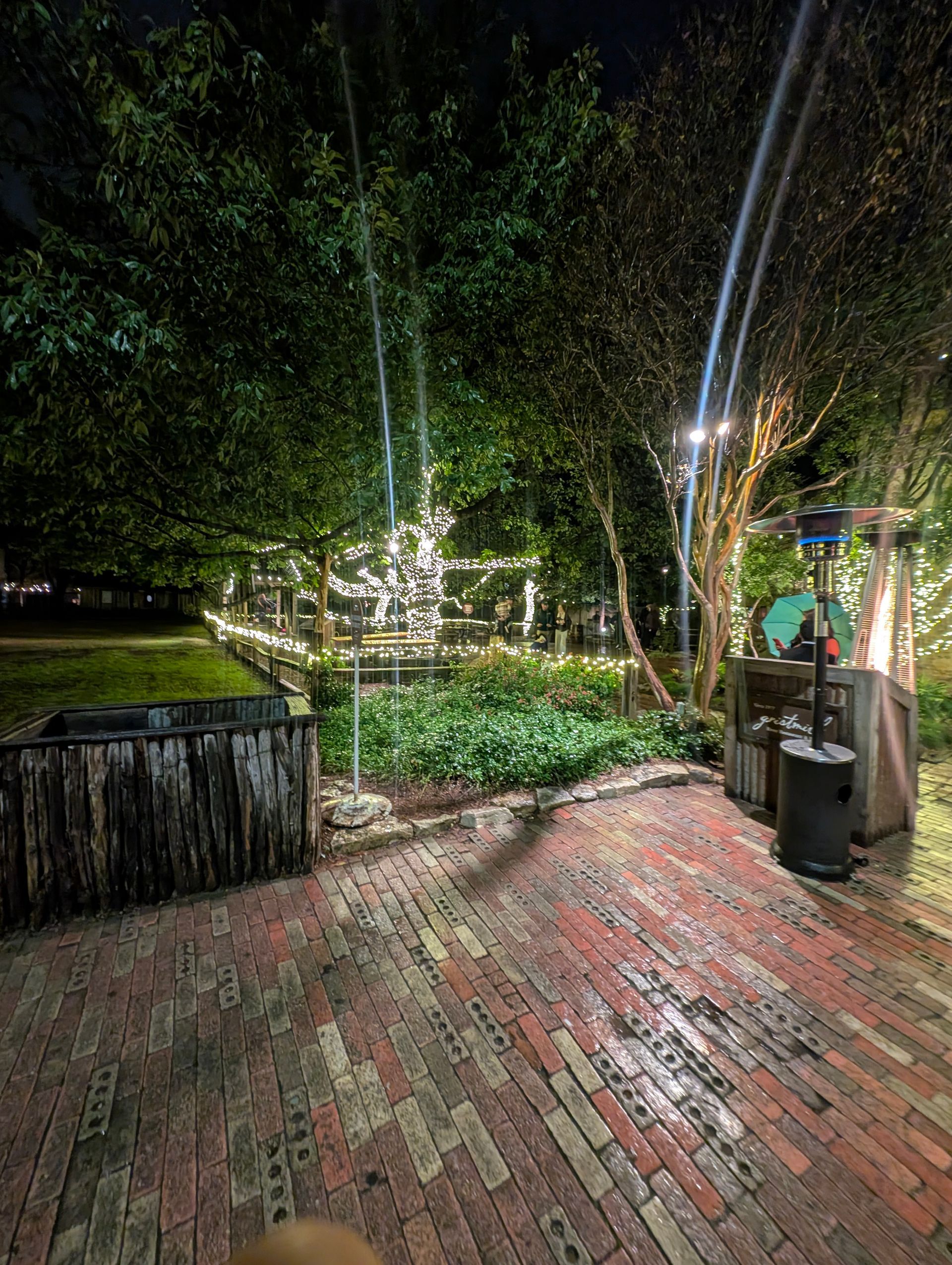 Brick patio at night with string lights in a garden; a heater and wooden trash can.