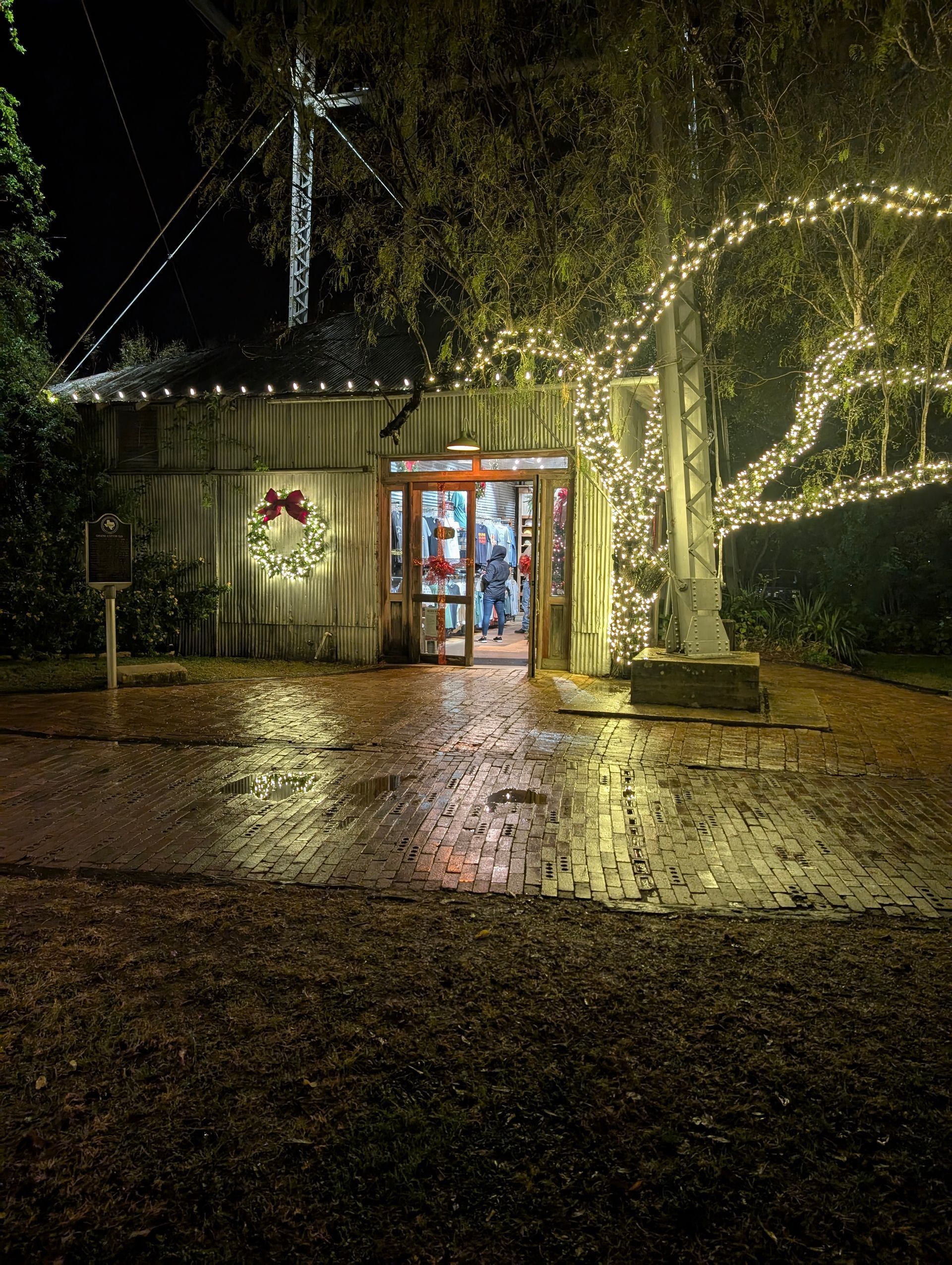 Night scene: Rustic building entrance, Christmas wreath, tree wrapped in lights, wet brick path.