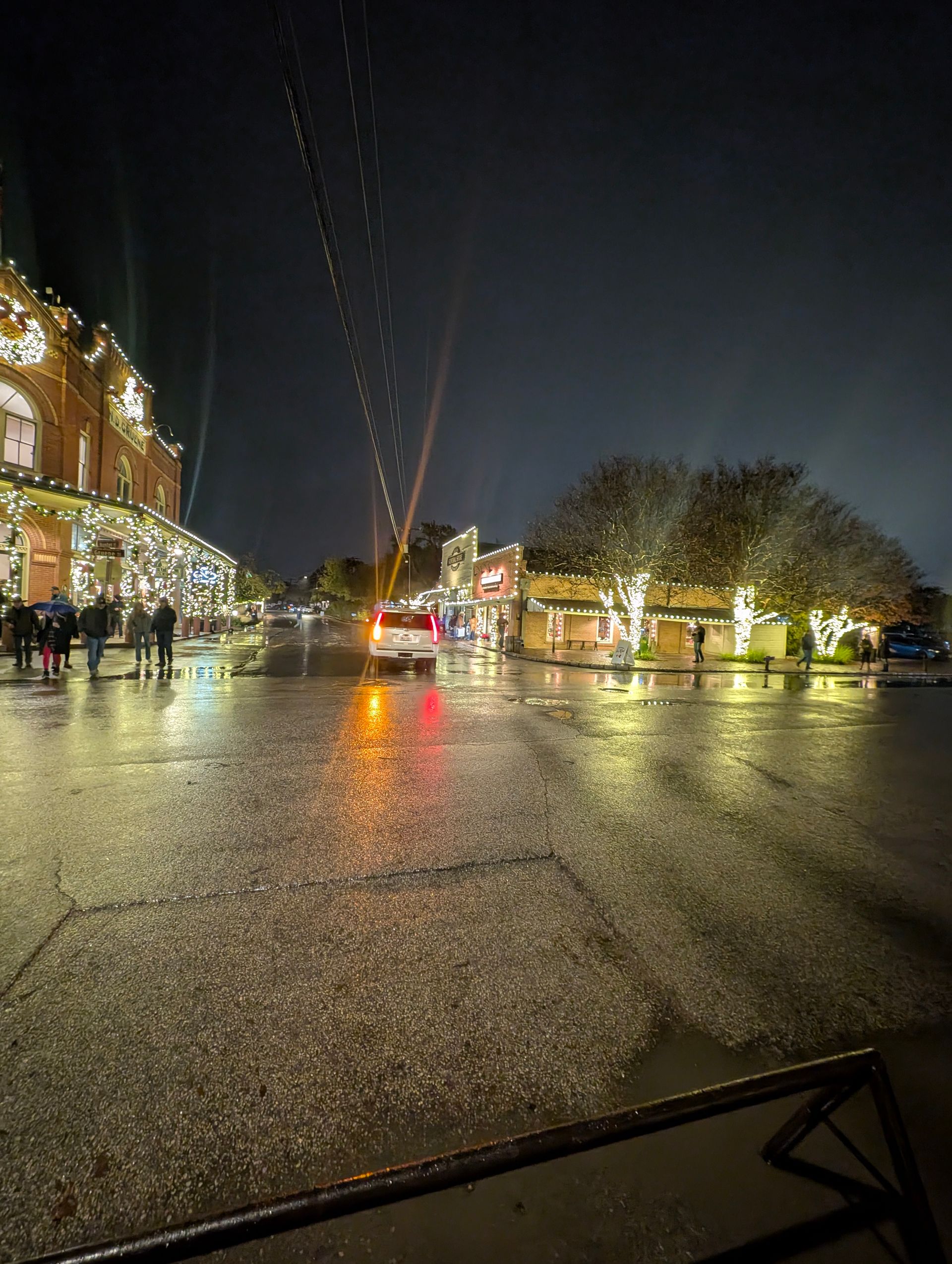 Wet street reflecting lights at night, decorated buildings with people.