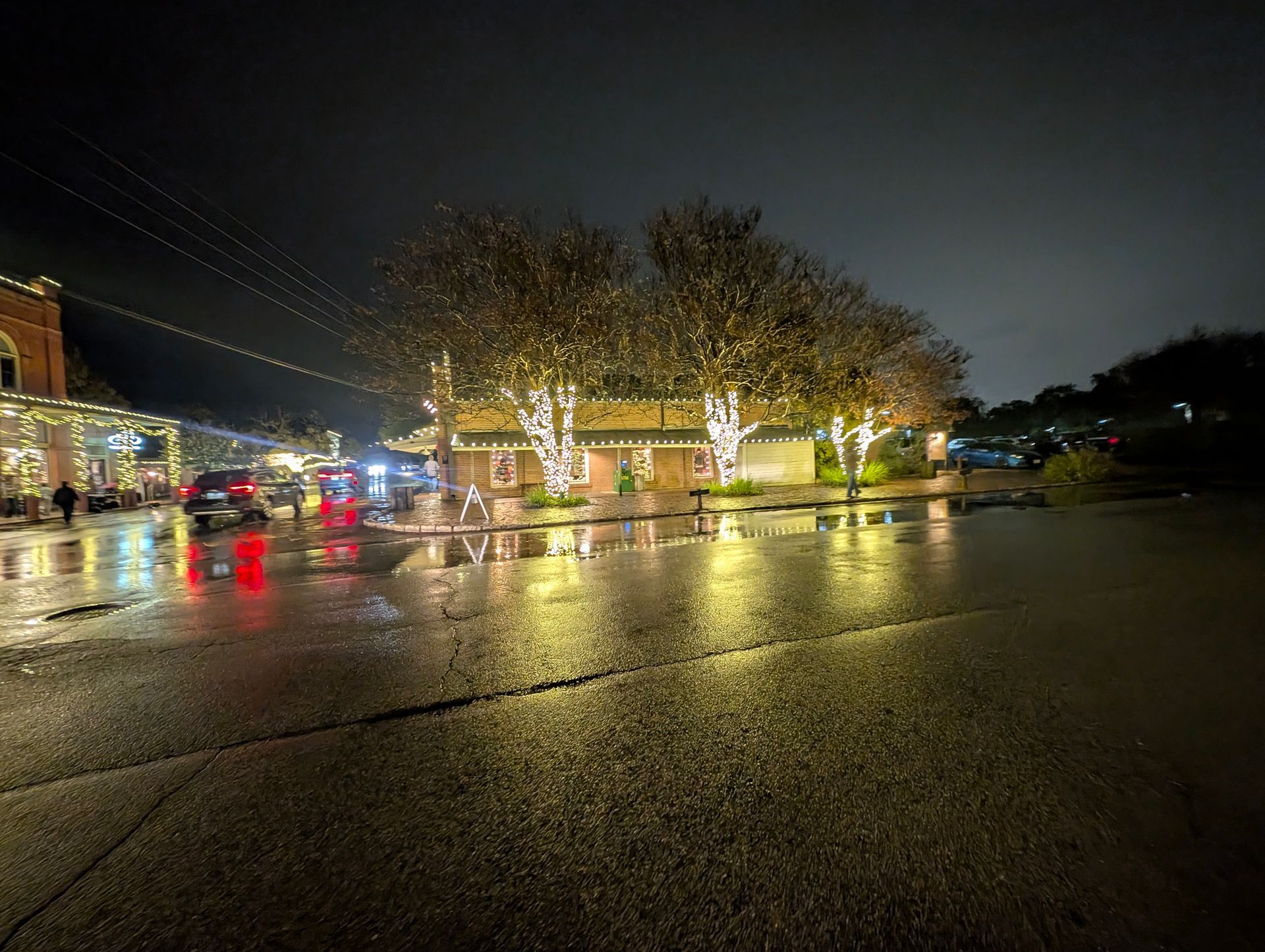 Wet street at night, lit by Christmas lights on buildings and trees, reflecting in the puddles.