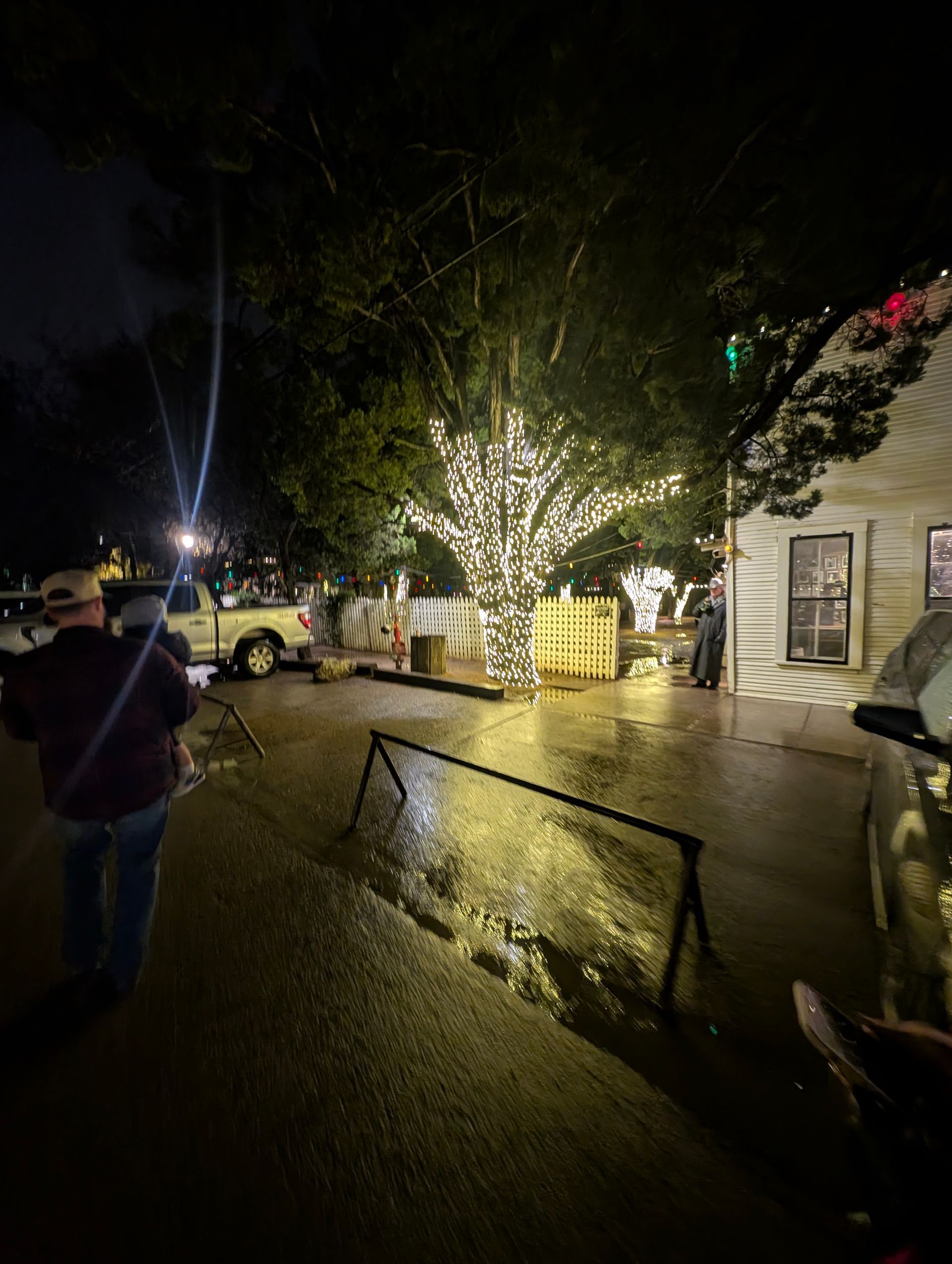 A night scene shows a tree lit with white lights, a white building, and people near a truck.