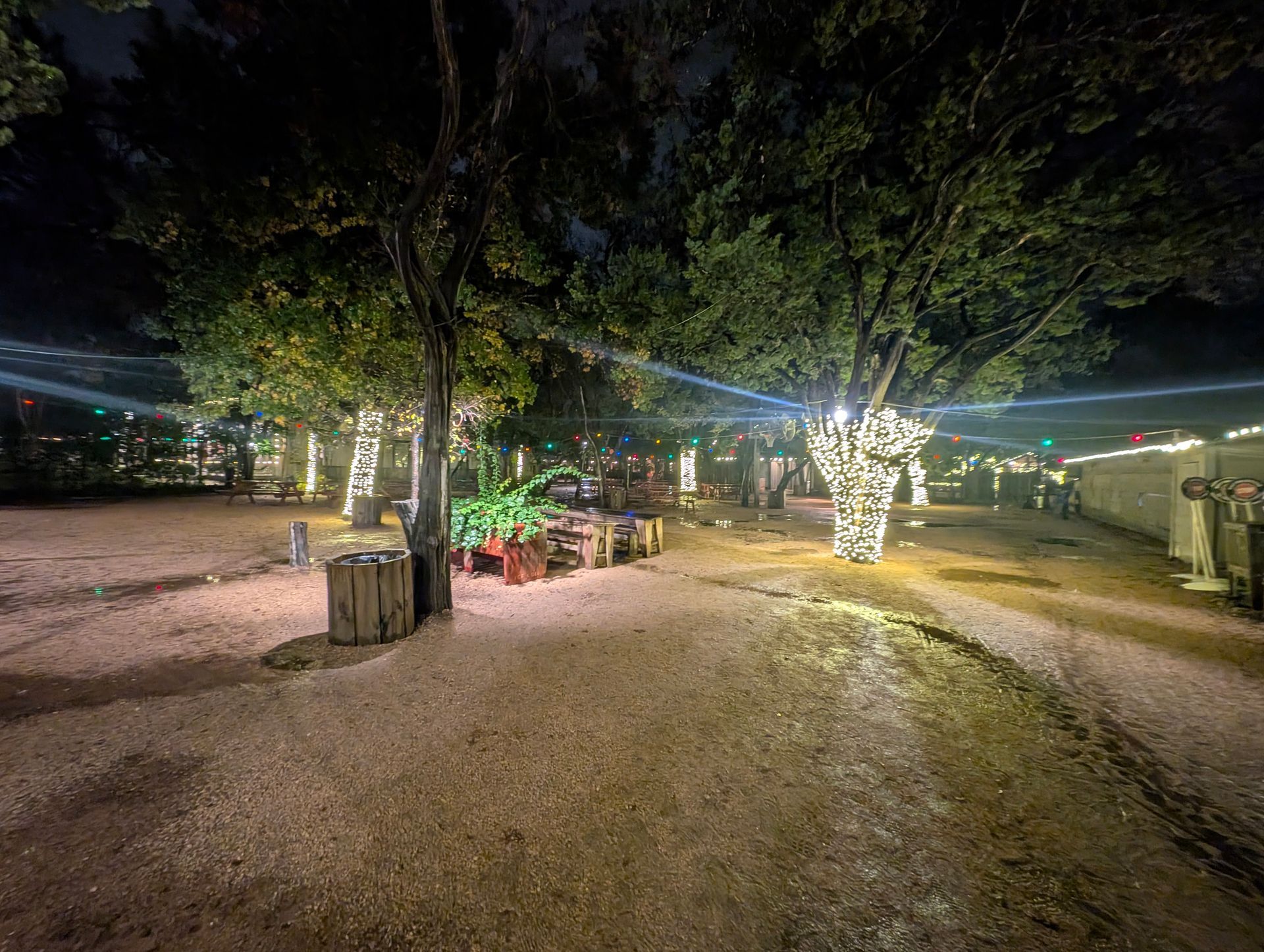Nighttime outdoor dining area; trees wrapped in lights, tables, and gravel ground.
