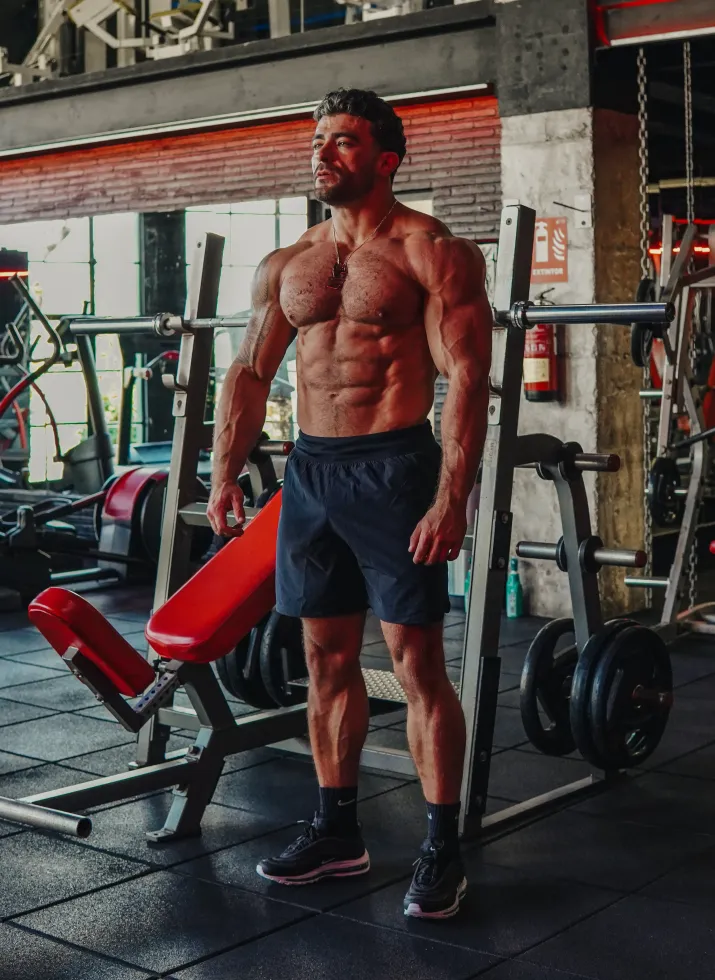 Jonny stands in gym, shirtless, by a bench press. Dark shorts, black shoes.