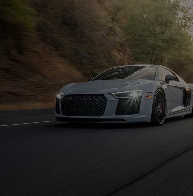 Gray sports car driving on a road next to a hill, headlights on | Balkan Auto Care