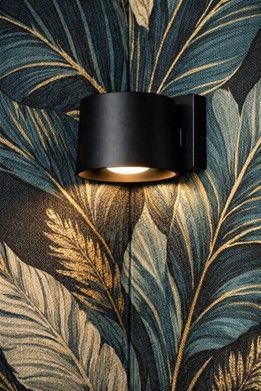 A matte black cylindrical wall lamp mounted against dark wallpaper featuring a pattern of green and gold tropical leaves.