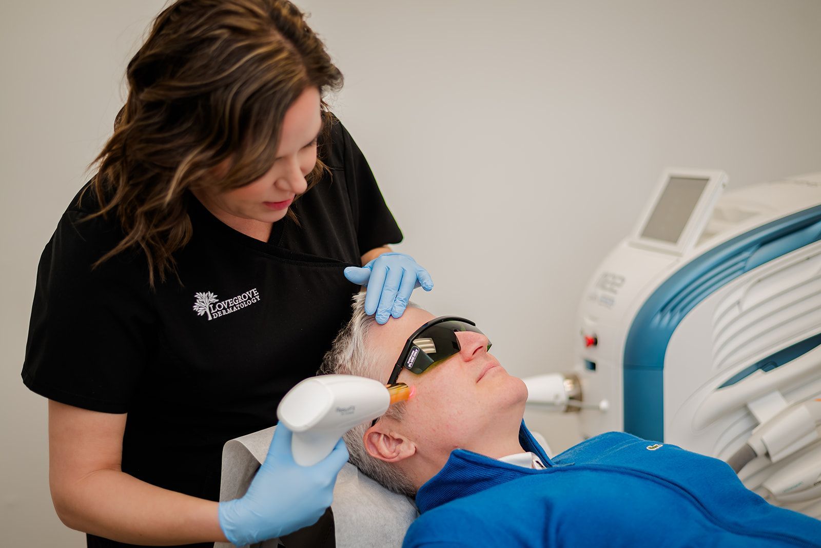 Medical aesthetician performs laser skin treatment on a man's face. Both wear safety glasses and gloves in a clinic setting.