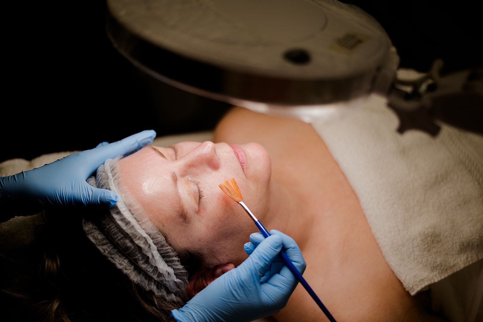 Woman receiving a facial chemical peel, being treated with a brush, under a magnifying lamp.