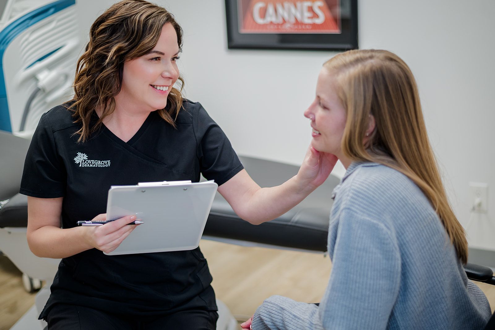 Medical aesthetician examines a patient's face, holding a clipboard in an office setting.