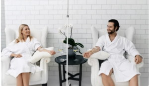Couple in white robes at a spa, sitting in armchairs, smiling, with drinks.