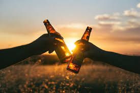 Two hands toasting beer bottles against a sunset backdrop.