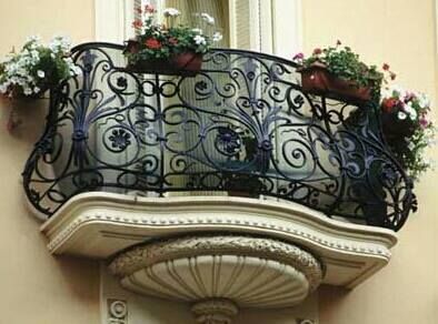 Balcone decorato in ferro battuto con fioriere, incastonato in un edificio dai colori chiari.