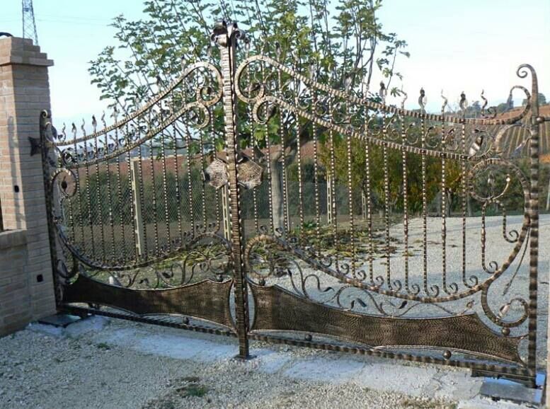 Cancello decorato in ferro battuto, bronzo e nero, di fronte a un vialetto di ghiaia, con pilastri in mattoni ai lati.