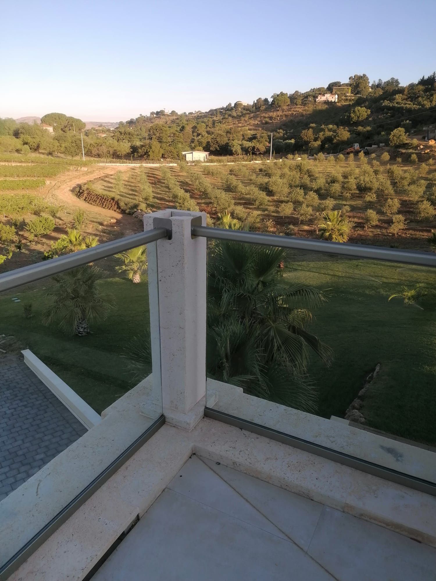 Vista da un balcone con ringhiera in vetro che si affaccia su campi verdi e colline sotto un cielo limpido.