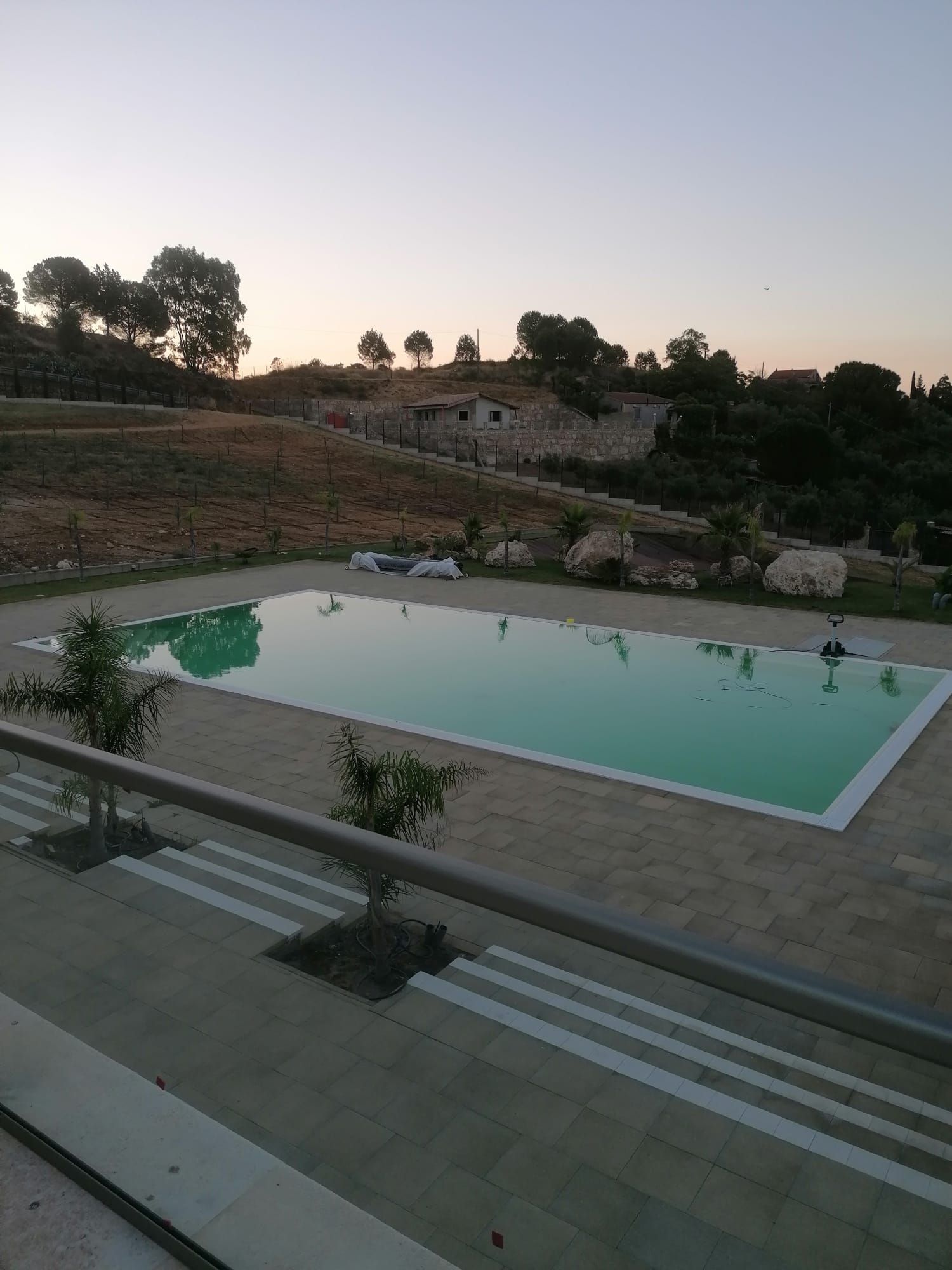 Piscina rettangolare in un ambiente rurale, circondata da un patio in pietra, con vista su un campo alberato, sotto un cielo al tramonto.