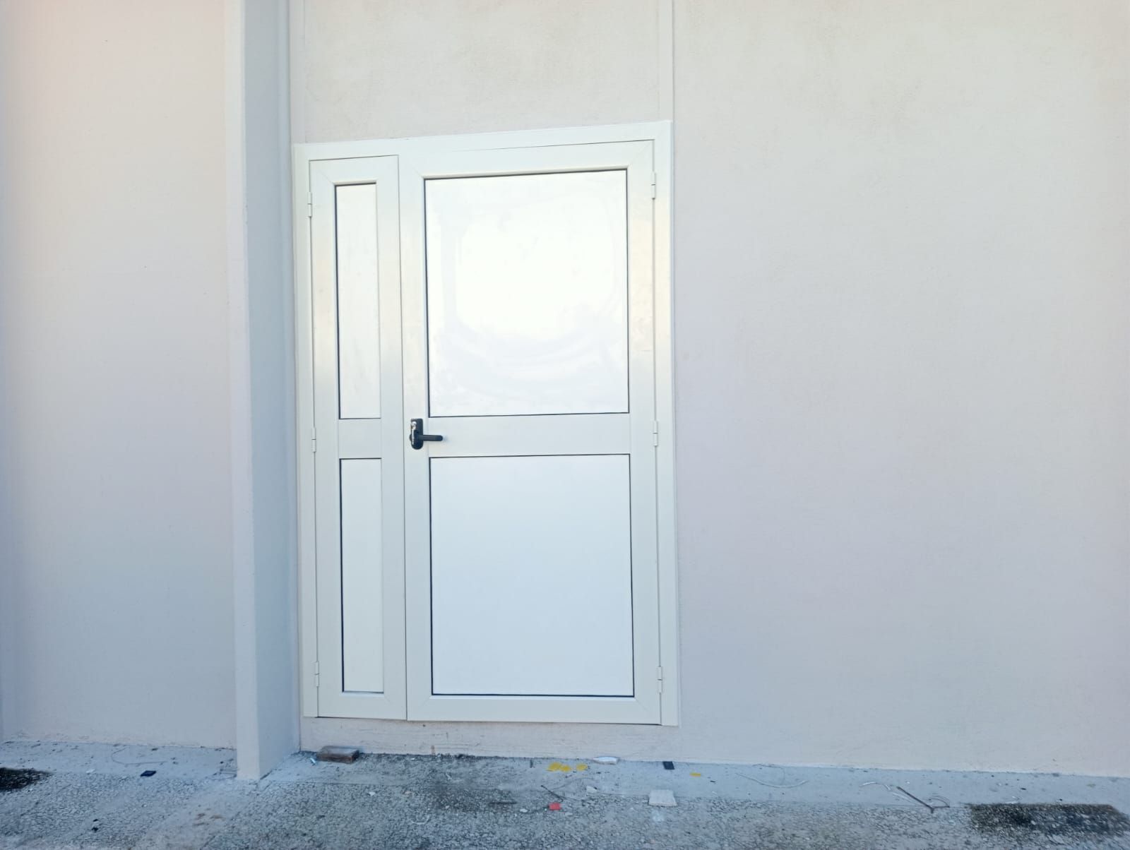 Porta bianca con pannello in vetro e finestra laterale inserita in una parete grigio chiaro.