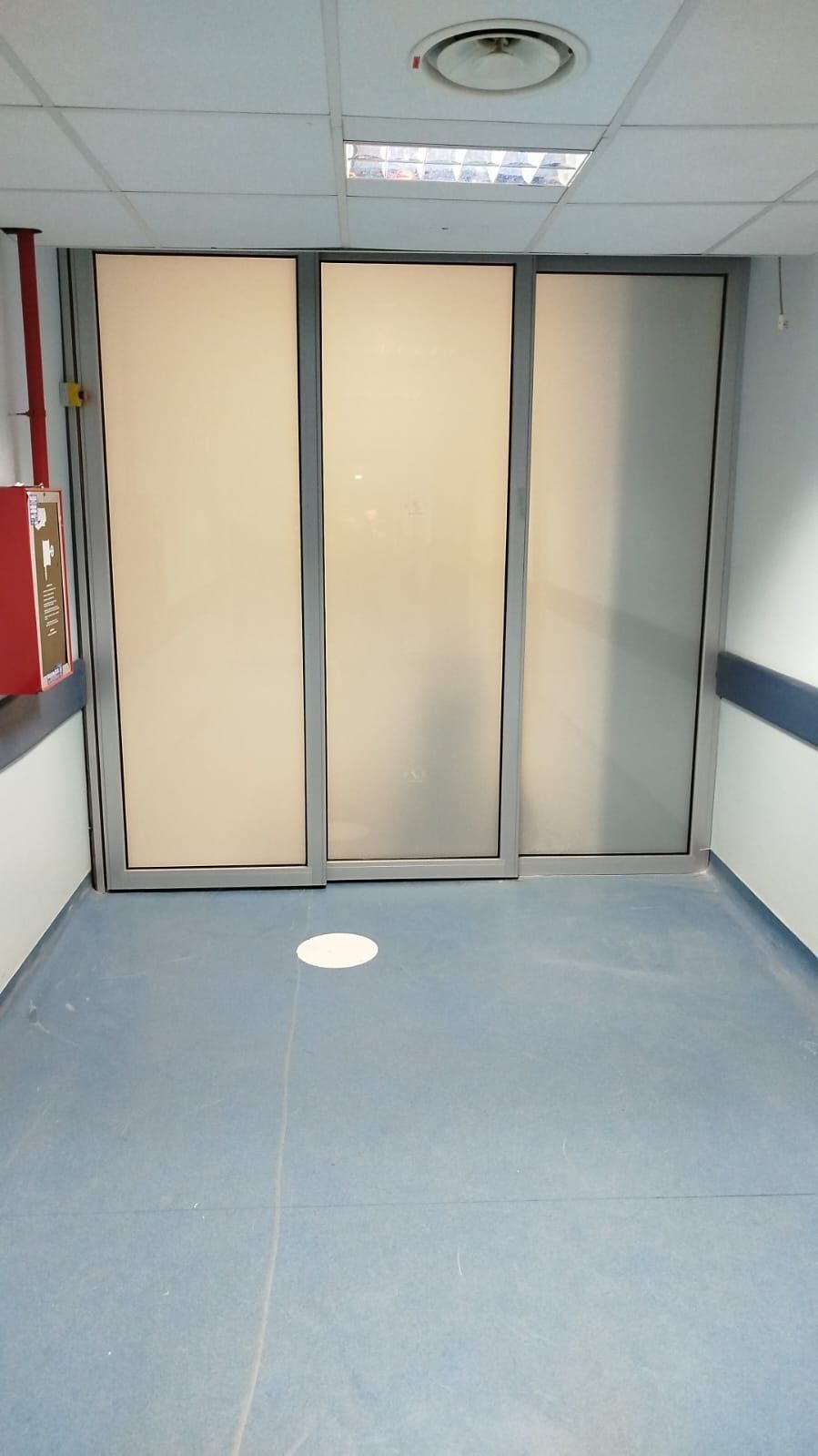 Porte in vetro smerigliato in un corridoio con pavimento blu, probabilmente un ospedale o una struttura medica.
