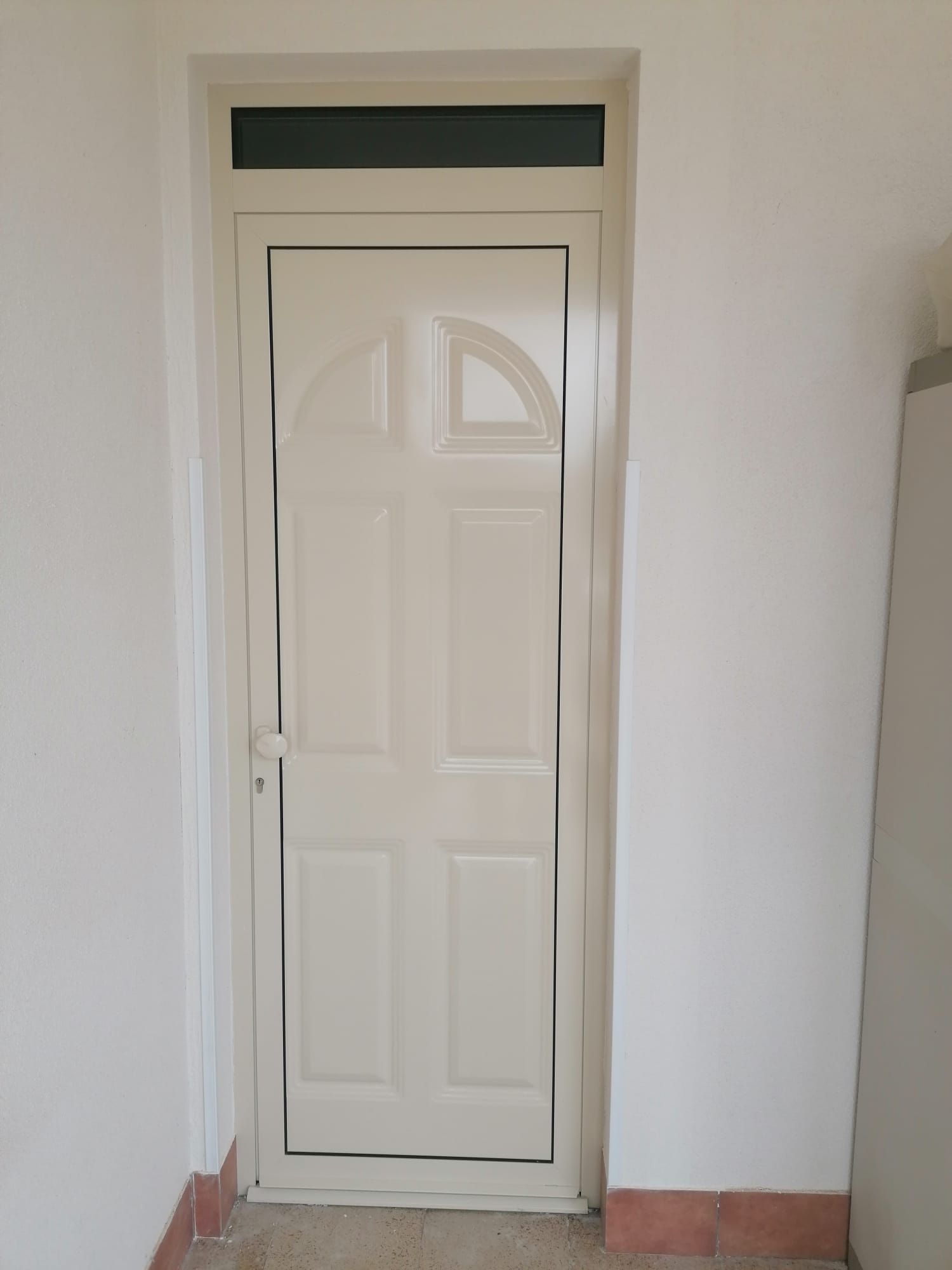 Porta bianca con pannello superiore ad arco e finestra a traverso, inserita in una parete bianca con pavimento in piastrelle rosse.