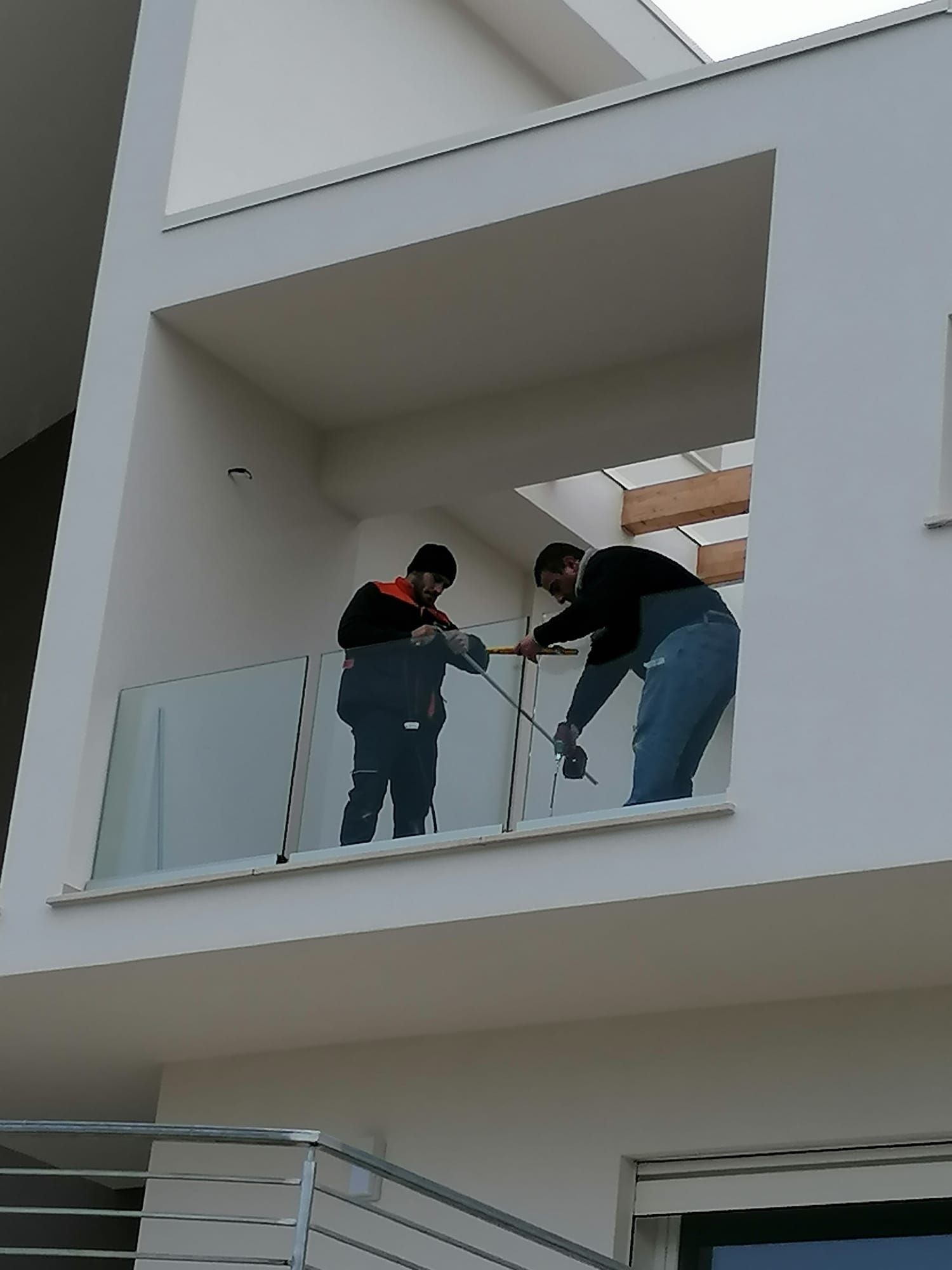 Due operai su un balcone stanno utilizzando degli attrezzi sulla ringhiera di vetro di un edificio bianco.