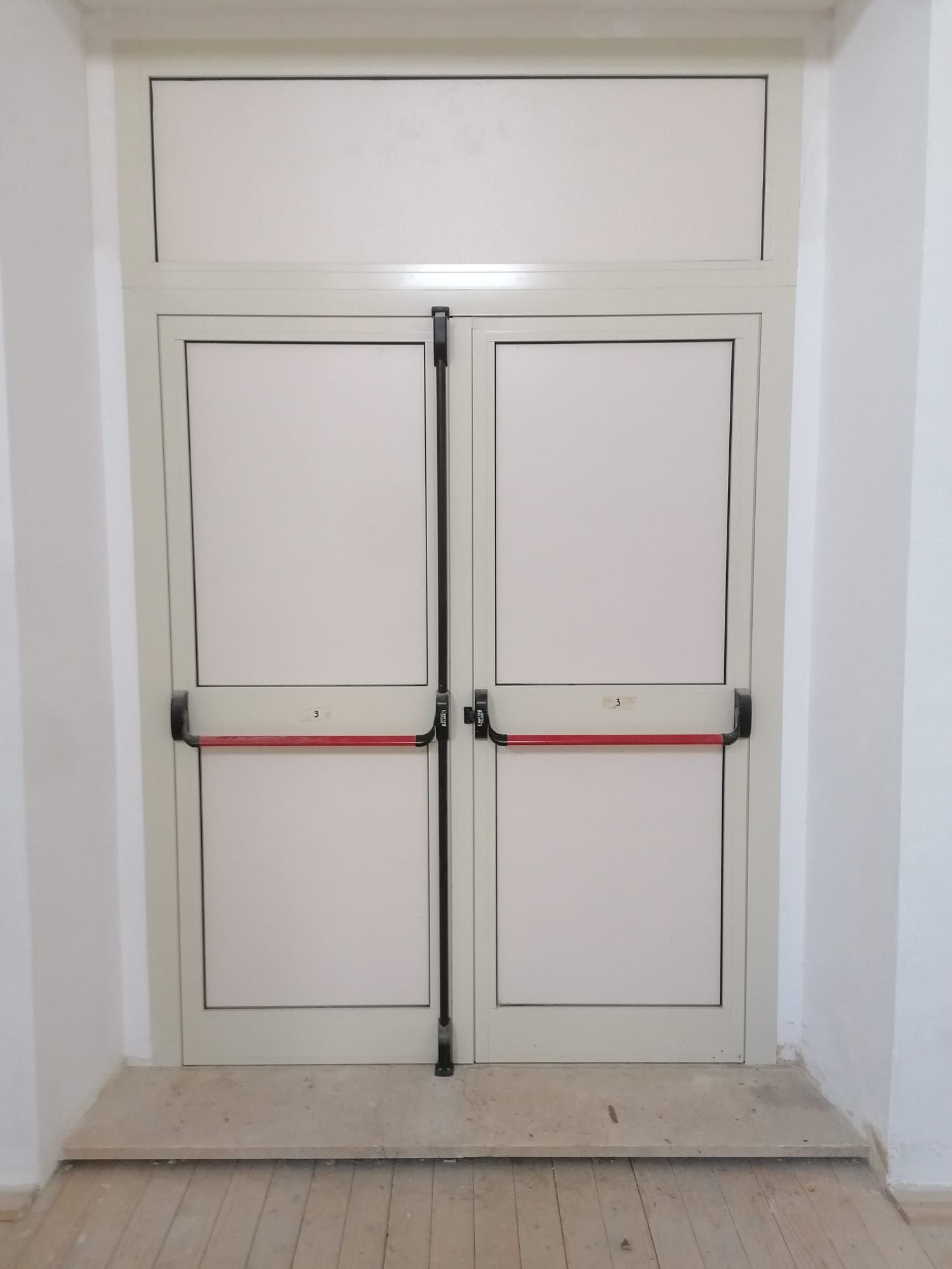 Porte doppie con finestra a traverso superiore. Maniglie di spinta rosse, telai beige, pannelli bianchi.