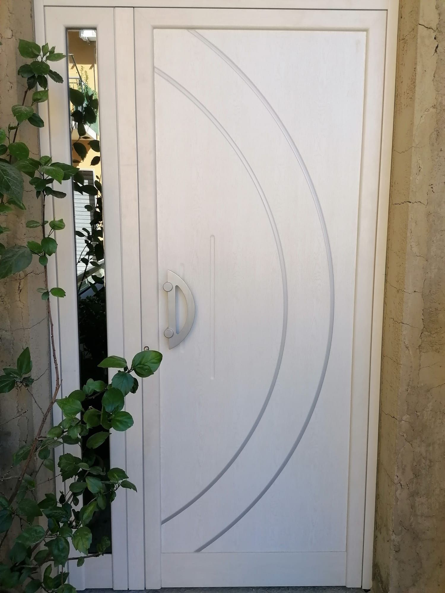 Porta d'ingresso bianca con linee curve e luce laterale, fogliame sulla sinistra, accanto a una parete color cuoio.