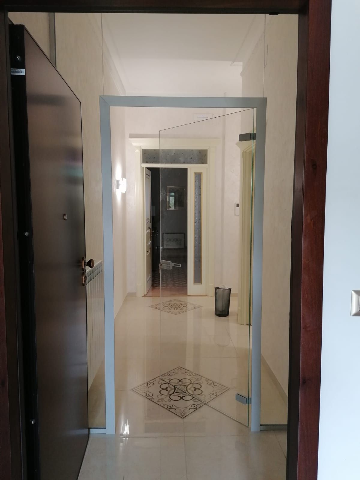 Corridoio interno con porta d'ingresso aperta. Una porta a vetri conduce a uno stretto corridoio con pavimento piastrellato e una porta in fondo.