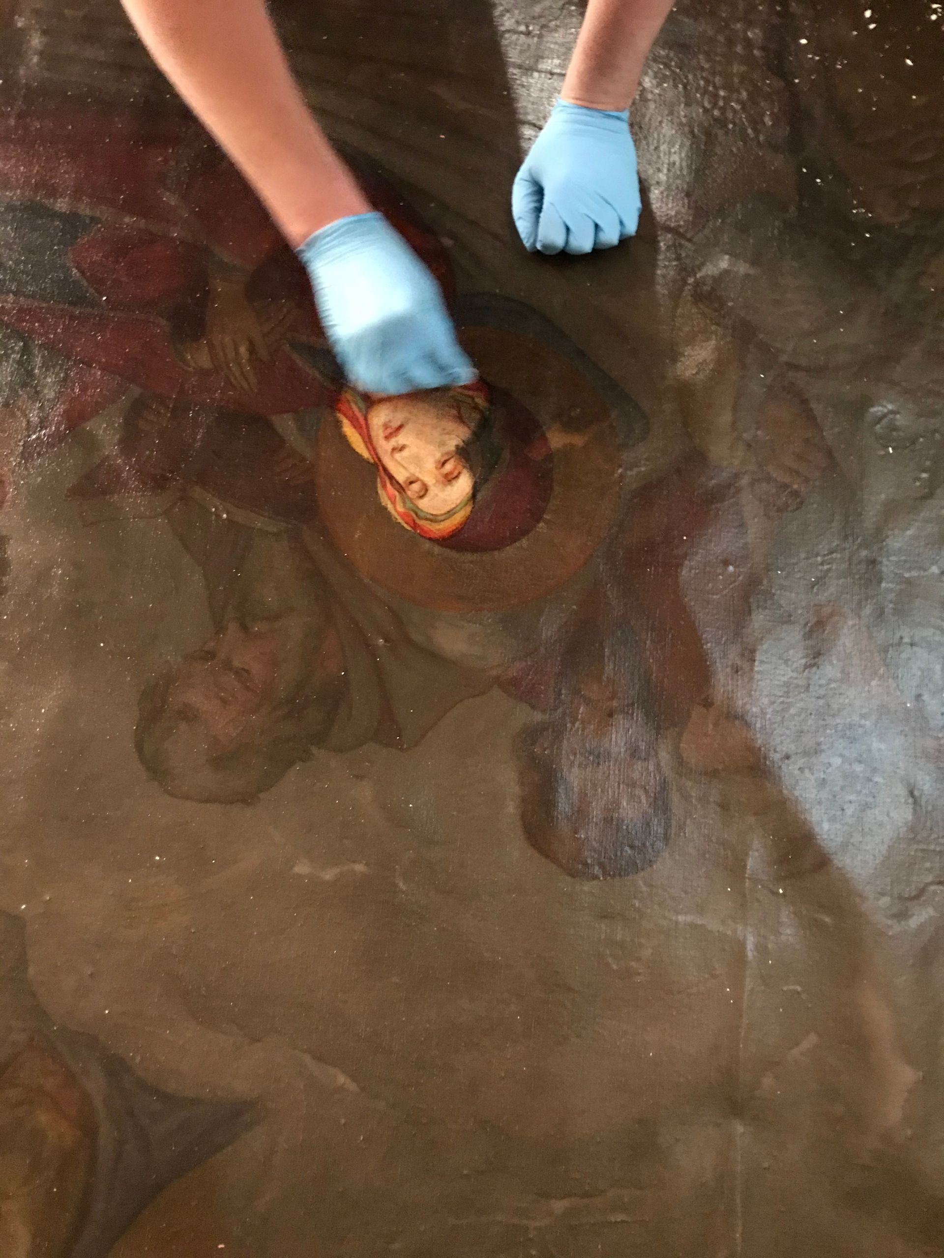 A person wearing blue gloves is cleaning a painting on the floor.