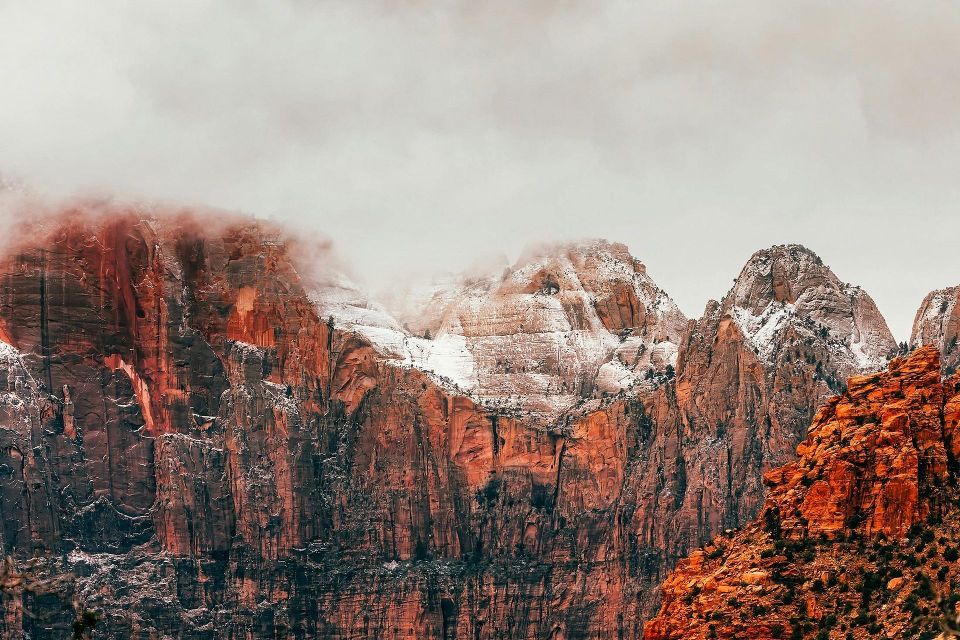 Red and tan rock mountains with snow-capped peaks under a cloudy, overcast sky.