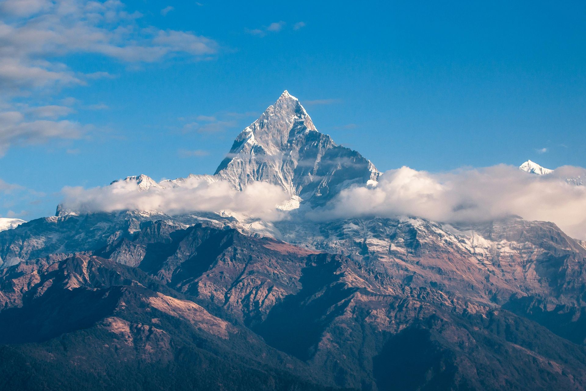 Snow-capped mountain peak piercing blue sky, nestled amongst brown and green ridges; wisps of clouds surround the base.