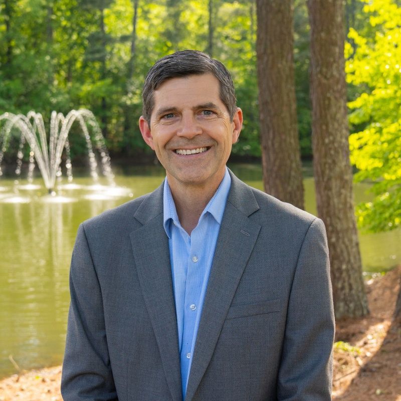 Man in a gray suit smiles outdoors, in front of a pond and fountain.