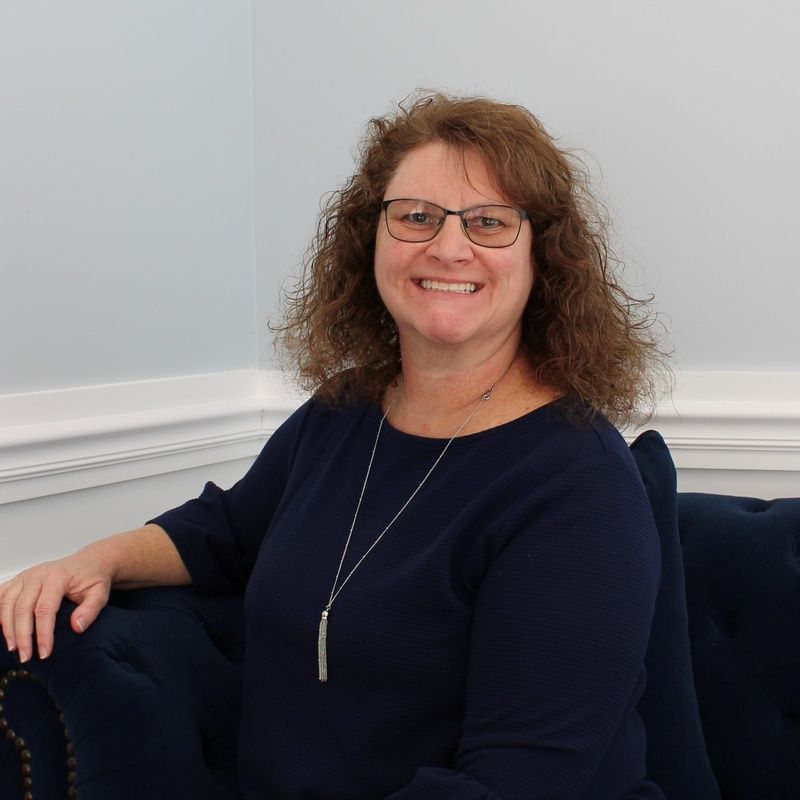 Woman with glasses smiles, sitting on a blue couch, wearing a navy top and necklace.