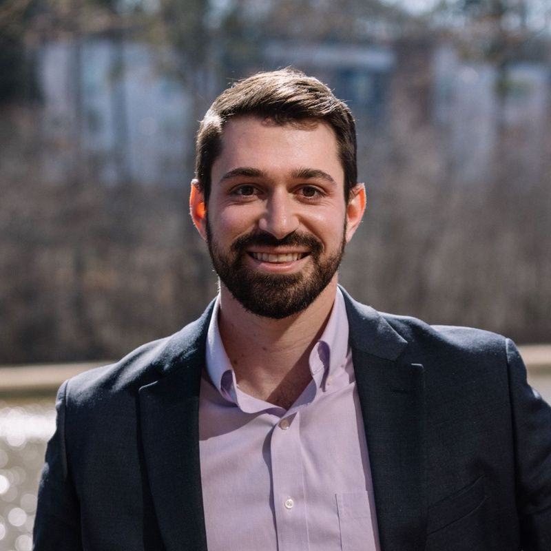 Man with a beard wearing a suit jacket and purple shirt, smiling outdoors.