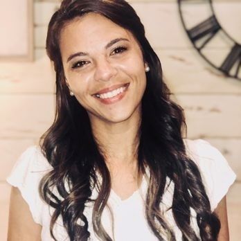 Woman with long, wavy brown hair smiles. She wears a white blouse and small earrings, indoors.