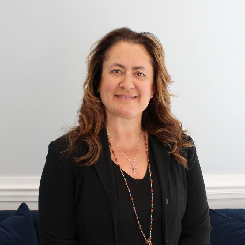 Woman with brown hair and a black blazer, smiling, wearing a necklace with a cross.